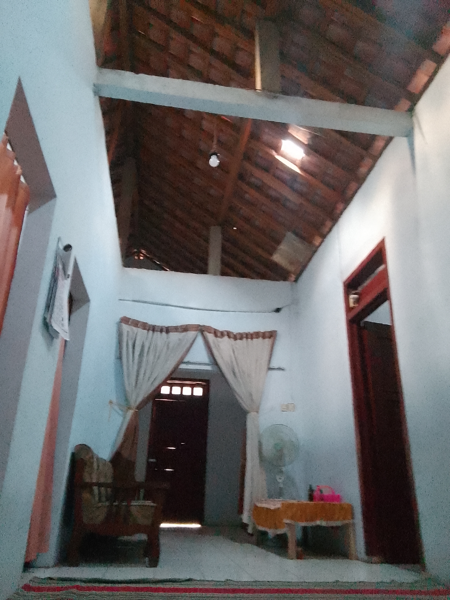 The image depicts an indoor scene, likely a living room or hallway, within a building located in Karangan, Indonesia. The room has a light blue, textured wall and a wooden-tiled ceiling with exposed beams. A prominent feature is a wooden-framed armchair with patterned upholstery situated on the left side of the frame. In the background, a dark wooden door with glass panes is visible, partially obscured by light-colored curtains. To the right of the door, a dark red door stands vertically. In the mid-ground, a small table with an orange tablecloth is present, topped with a pink object, possibly a basket or bag, and a black bottle. A standing fan is positioned behind the table. The floor is covered with a rug featuring red and green stripes. The overall lighting suggests it is daytime, with light entering from a window or opening in the ceiling.