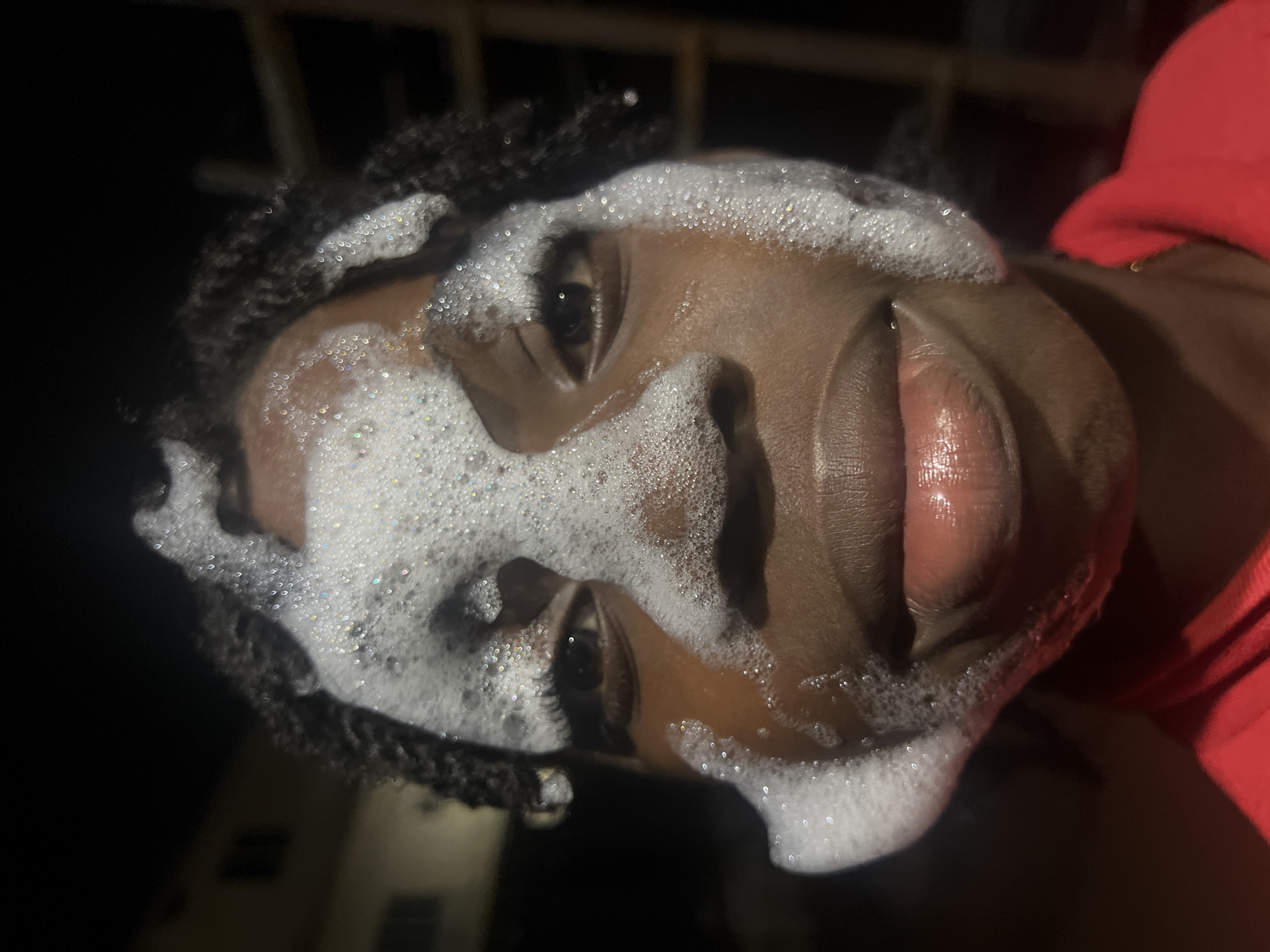 A close-up, eye-level, waist-up shot captures a person with dark skin and braided hair in Nkpor, Nigeria, at night. The individual's face is partially covered in white foam, suggesting a bath or a playful moment. The foam obscures the forehead and extends down the cheeks and around the nose. Their eyes are visible, looking directly at the camera with a neutral expression. The lips are slightly parted, also coated with some foam, and appear glossy. The person is wearing a red shirt. The background is dark and out of focus, with hints of a building and possibly a ladder, indicating an outdoor or semi-outdoor setting in a residential area at night. The lighting is likely artificial, highlighting the foam and the person's face.