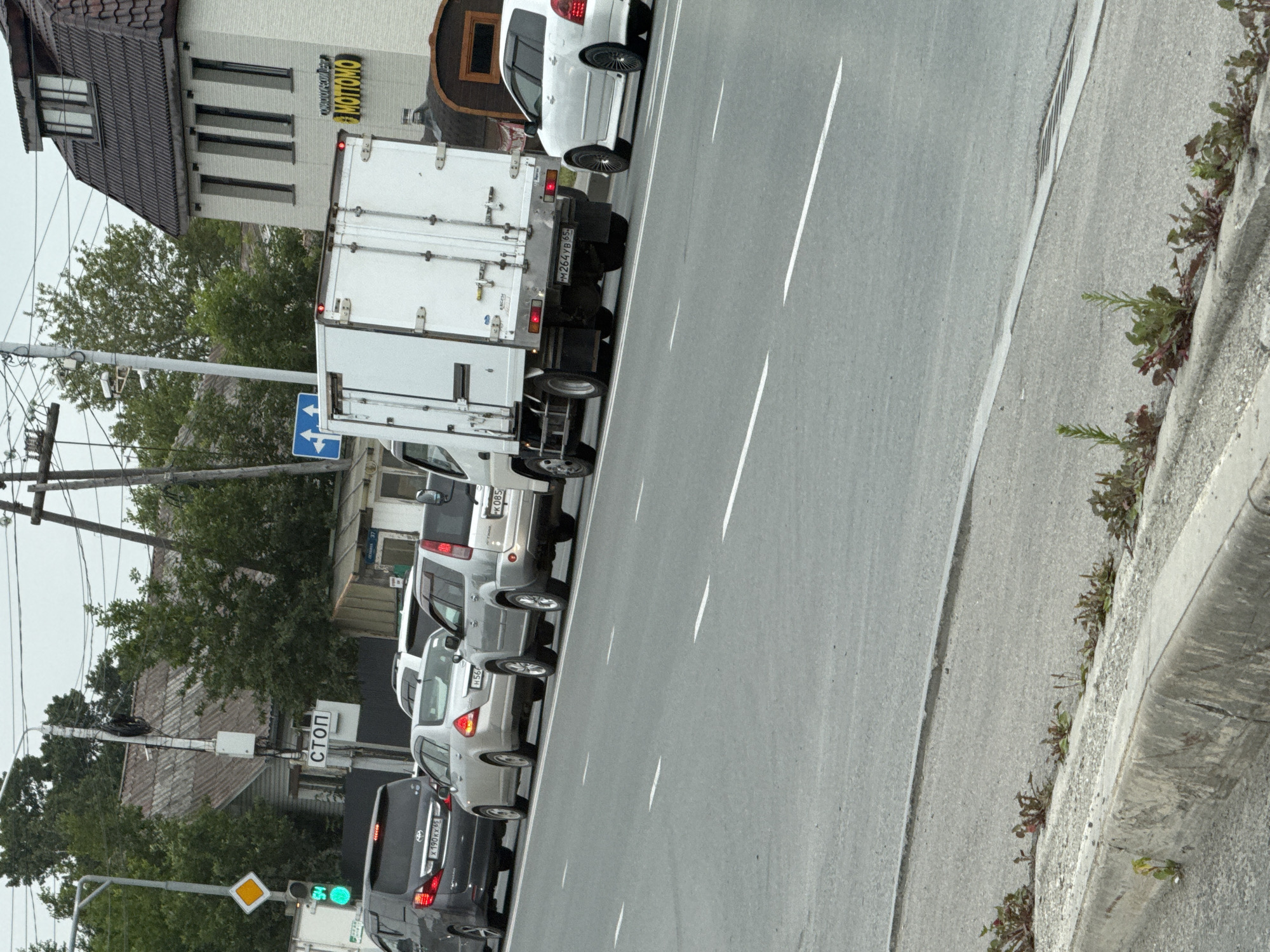 The image captures a traffic scene in Yuzhno-Sakhalinsk, Russia, on a slightly overcast day. The primary focus is a multi-lane road with several vehicles queuing. In the foreground, the asphalt road is visible with white lane markings. A concrete curb runs along the side of the road, with sparse vegetation growing in the cracks and on the shoulder.
Several cars are lined up on the road. A dark grey minivan, a silver sedan, a silver SUV, and a white truck are prominently featured. A white sedan is also visible further down the road. A sign with the Russian word "СТОП" (STOP) is mounted on a pole on the left side of the image, indicating an intersection or traffic control point. A traffic light is also visible, showing a green light.
In the background, there are buildings and trees. A multi-story building with a dark tiled roof and white walls is visible on the right, with a sign that appears to read "MOTTOМО". Further left, a residential building with a grey, corrugated roof is seen behind the traffic signs and trees. Overhead power lines are also present in the background. The overall impression is a typical urban street scene in Russia.