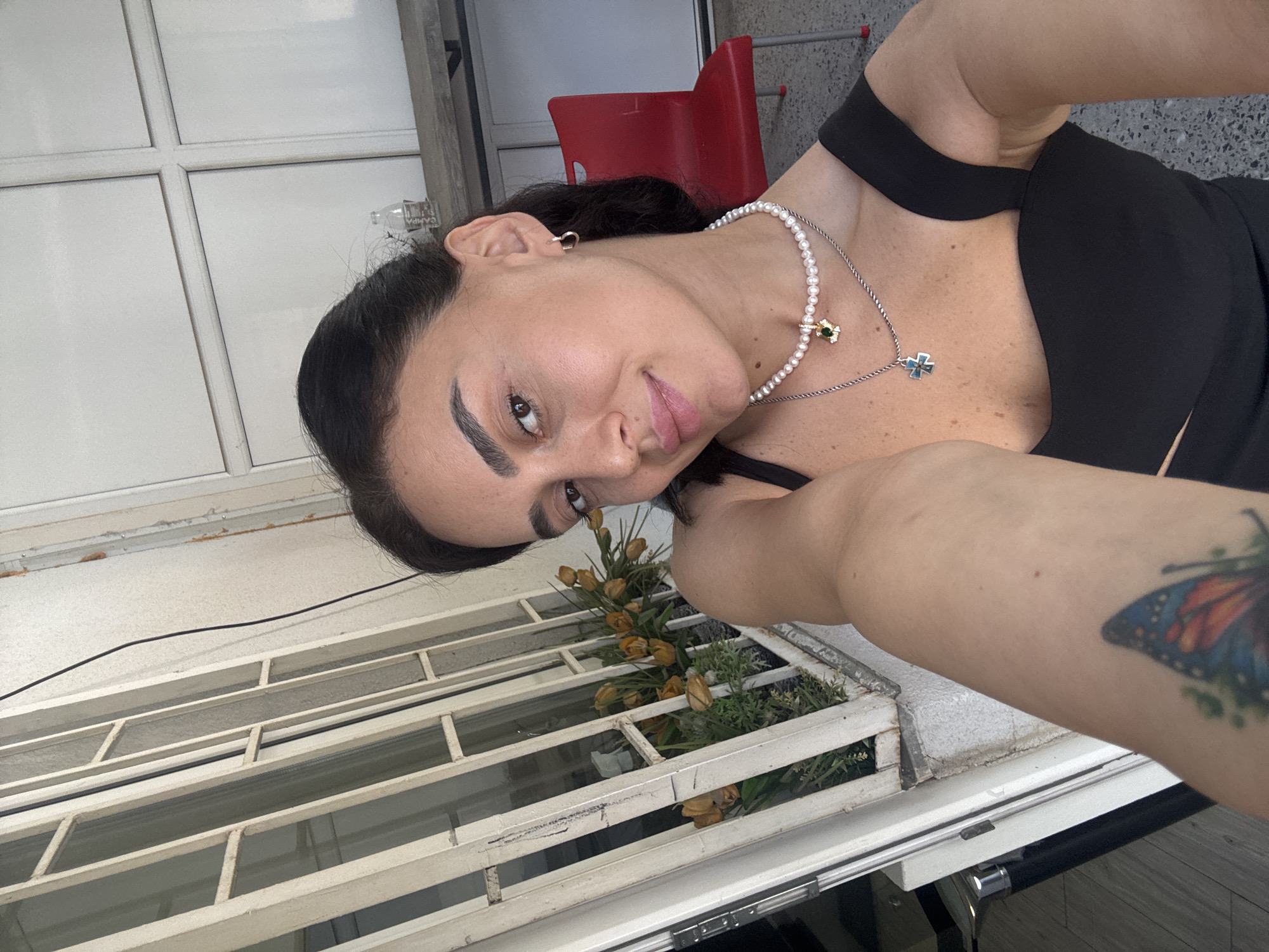 A woman with dark hair and fair skin is taking a selfie. She is wearing a black top and two necklaces, one with pearls and a green pendant, the other with a blue cross pendant. She has a butterfly tattoo on her left arm. The background shows a white metal grate, some dead flowers in a planter, a red chair, and a grey wall. This scene is consistent with an outdoor seating area or balcony in Tbilisi, Georgia.
