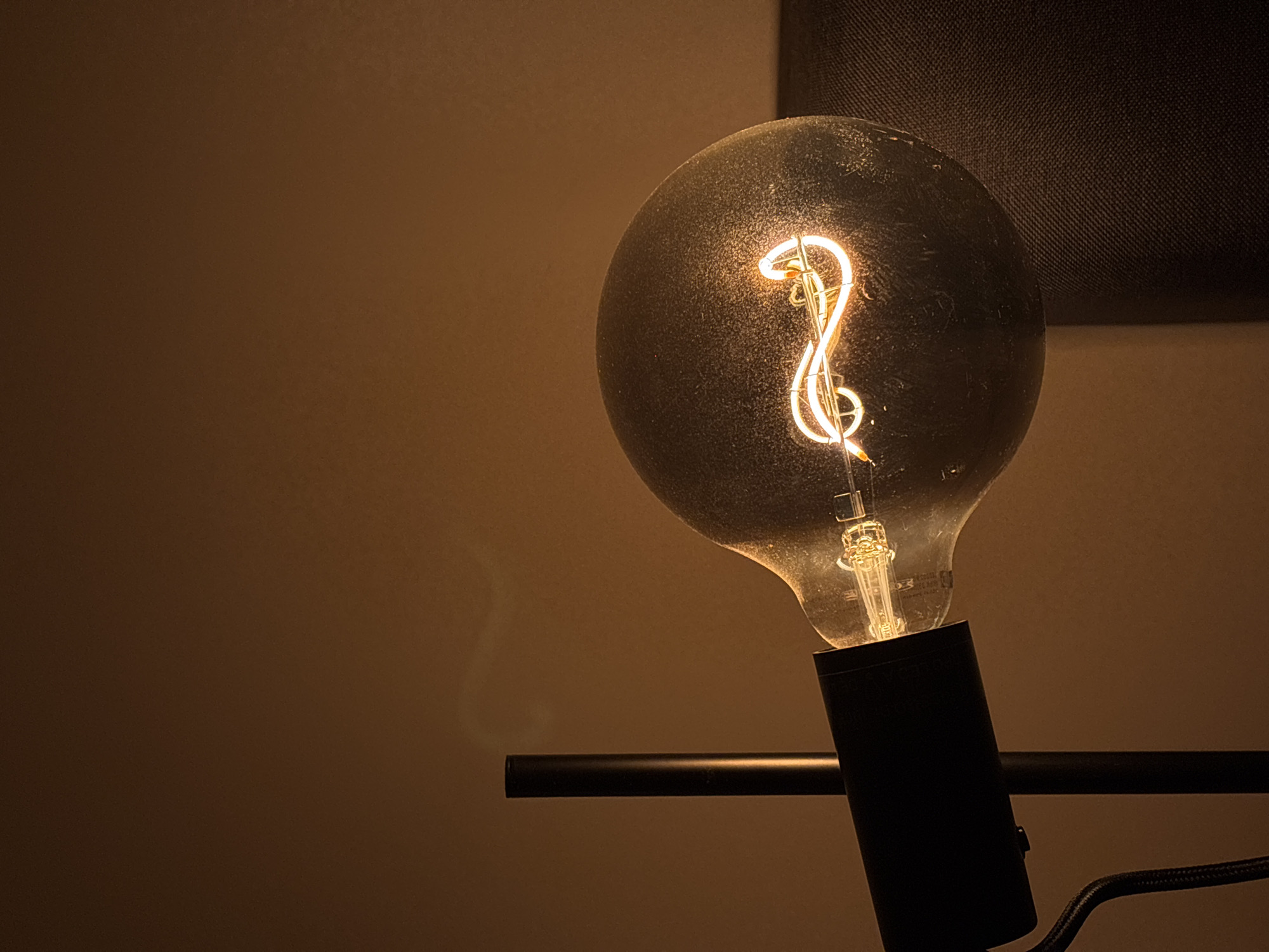 The image shows a lit lightbulb with a curly filament inside, mounted on a black fixture with a horizontal arm and a braided cord. The bulb is off-center to the right. The background is a warm brown wall on the left and a darker, textured surface, possibly fabric, in the upper right corner. The scene is dimly lit, creating a moody atmosphere. The location in Santa Clara Ocoyucan, Mexico is unknown and cannot be determined from the image.