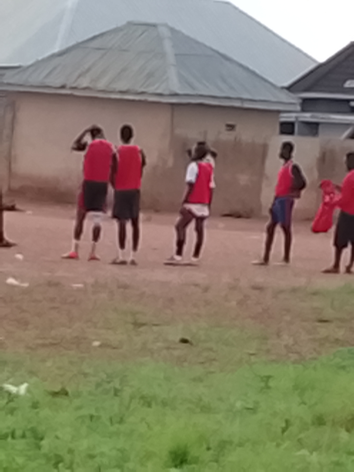 In Rafin Bauna, Nigeria, a group of young people, likely male, are gathered outdoors in front of simple, stucco buildings with corrugated metal roofs. The ground is dirt, with some patchy green vegetation in the foreground. Several individuals are wearing red vests over their clothing, and some appear to be engaged in a casual activity, possibly a sport or practice session. One person on the far right is holding up a red garment. The overall atmosphere is informal and suggests a community gathering or recreational event.