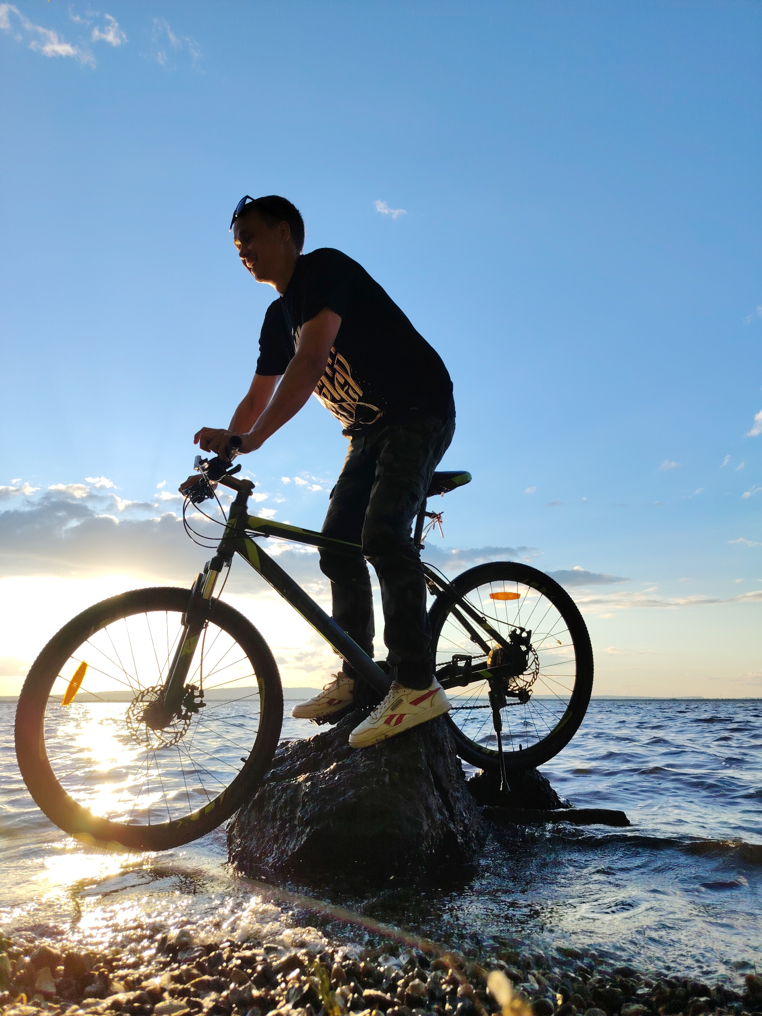 A young Asian male is seen from the side, silhouetted against a bright sky and water, riding a bicycle that is perched on a rock in the shallow water. He is wearing a black t-shirt, dark pants, and sneakers. The bicycle is dark colored with green accents, and it has disc brakes. The sun is low in the sky, casting a warm glow and reflecting on the water. The water is calm with small ripples, and there is a rocky shoreline in the foreground. The sky is blue with some scattered clouds. The image appears to be taken at sunset or sunrise. The location is the Kama River embankment in Chaikovsky, Russia, known for its scenic views and recreational opportunities.