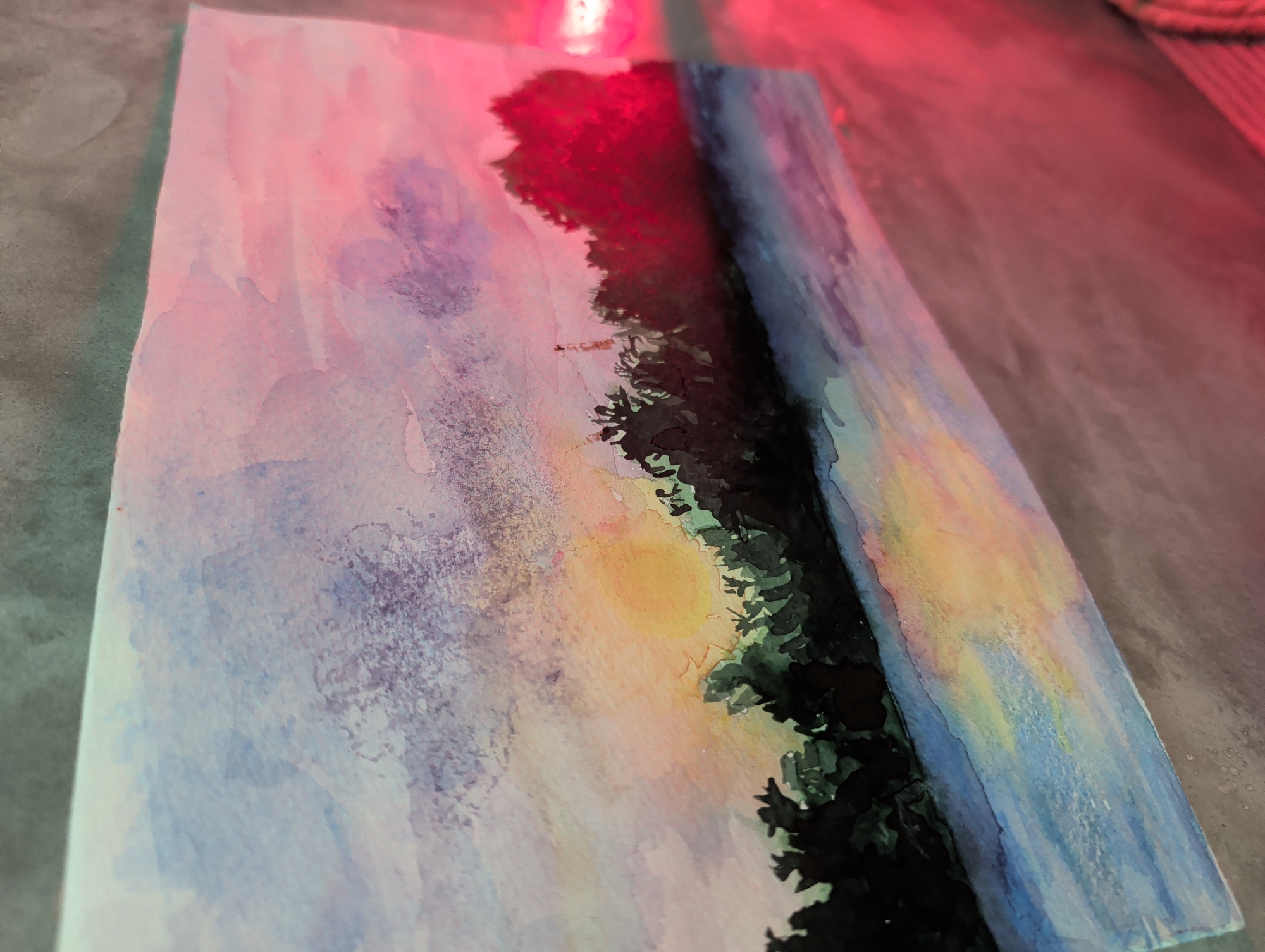 A watercolor painting depicting a landscape is displayed on a gray surface under red lighting. The painting features a sky with pink, purple, yellow, and blue hues, transitioning into darker shades near a line of trees. The trees are depicted as dark silhouettes, and in the center, there's a suggestion of sunlight. The painting's composition creates a sense of depth and perspective. The painting is angled slightly to the right. The scene depicts a nature scene in Ko Pha-ngan, Thailand.