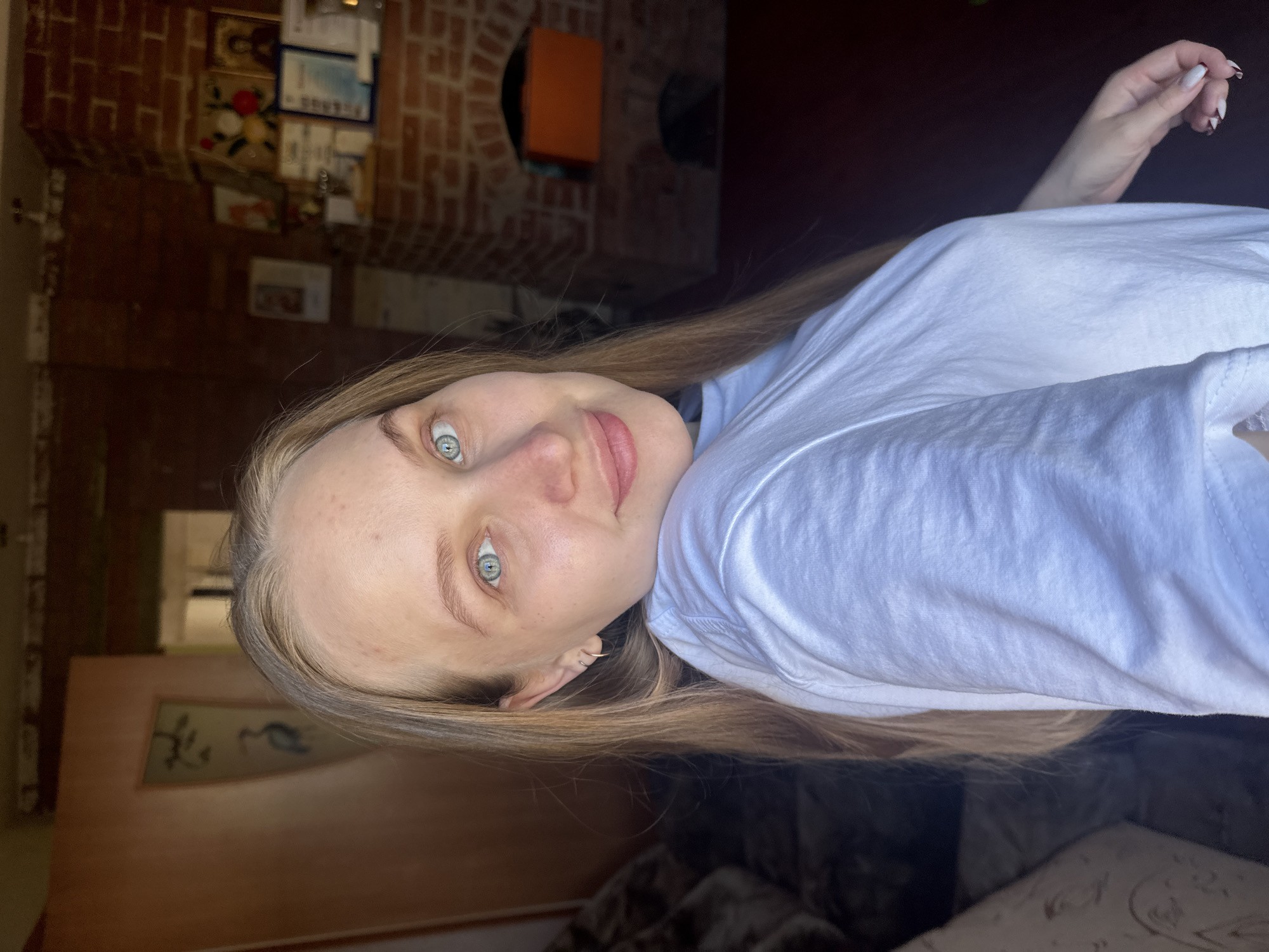 A young woman with long, light brown hair and light blue eyes is smiling at the camera. She is wearing a white t-shirt. The background shows the interior of a room with a brick fireplace and various decorative objects on the wall. The location is Chaikovsky, Russia.