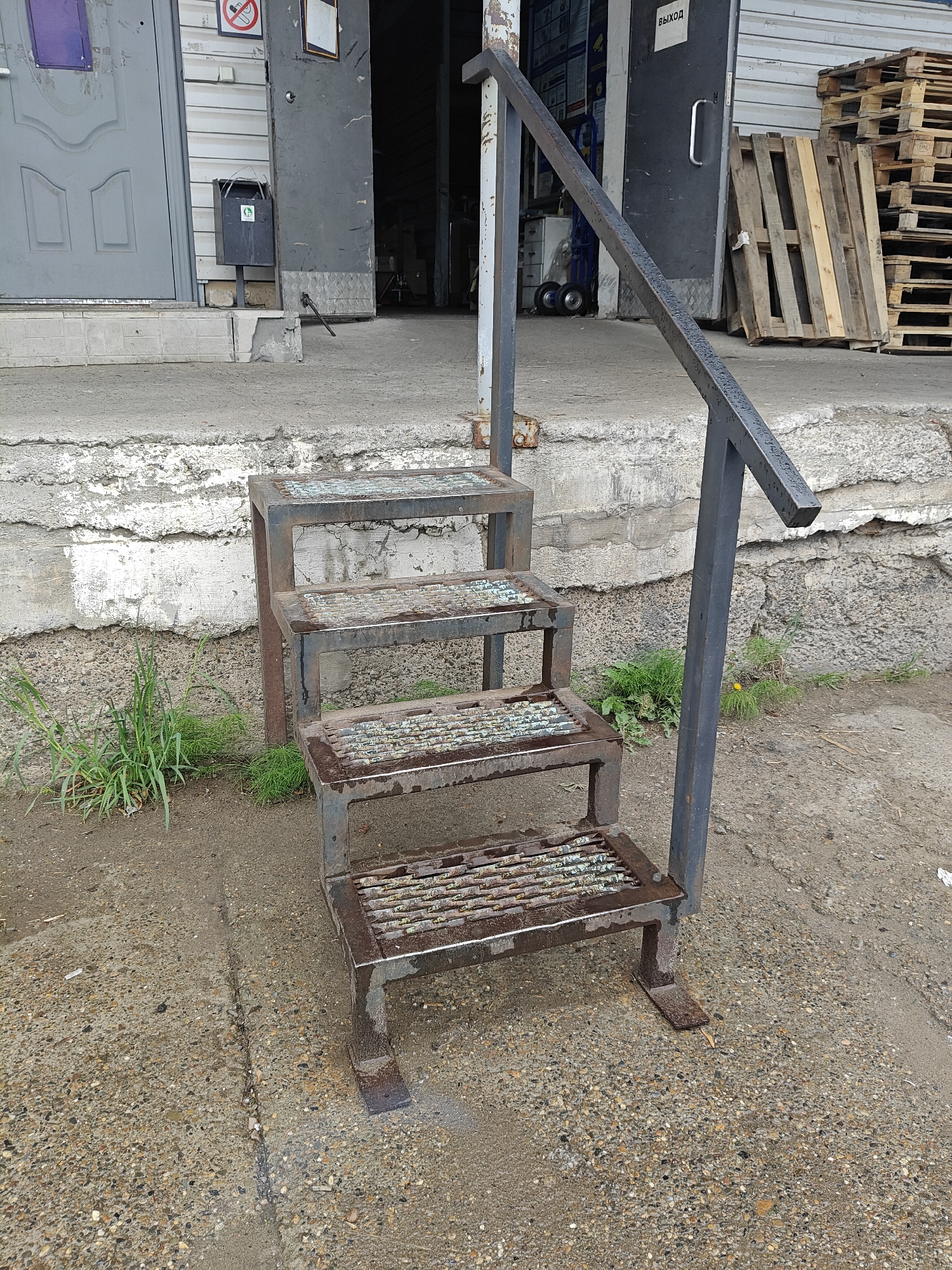 The image shows a set of metal steps with a handrail, positioned outside a building with a gray door labeled "ВЫХОД" (Exit) in what appears to be a loading dock area. The steps are rusty and weathered, suggesting they are old. The ground is concrete with cracks and some vegetation growing nearby. In the background, there are wooden pallets stacked against the wall and other objects are partially visible inside the open door. The city is specified as Chaikovsky, Russia.