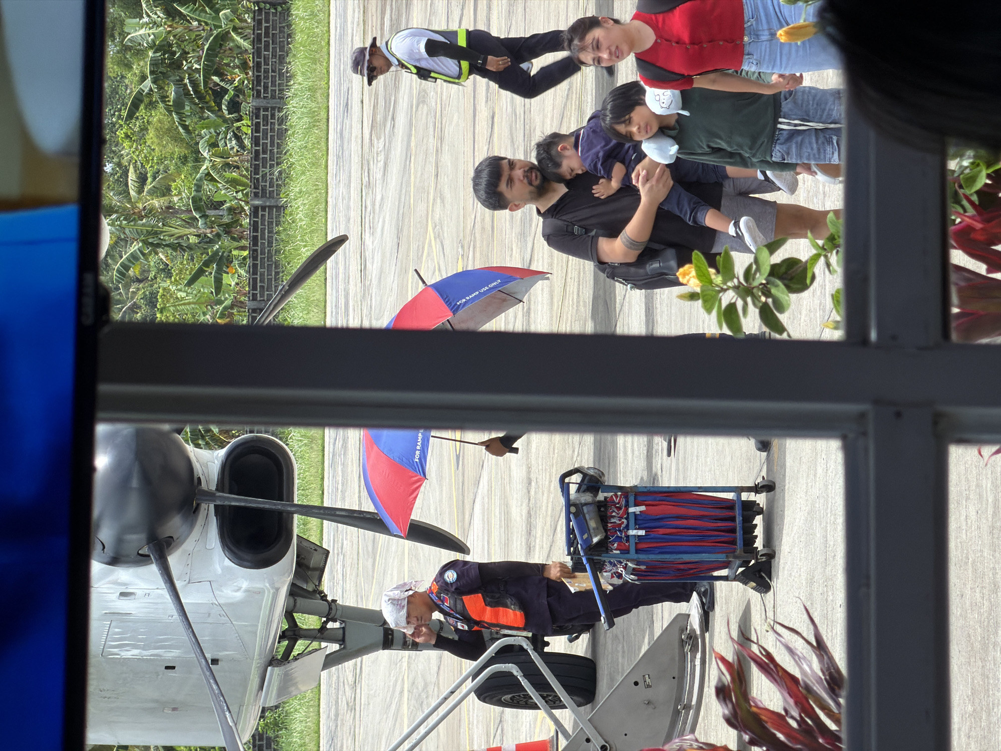 From an indoor vantage point, the scene unfolds at the Del Carmen airport in the Philippines. The foreground is dominated by a window frame. Outside, a plane and ground crew are visible. A man in a reflective vest stands with equipment, while another individual holds a red and blue umbrella. Adjacent to them, a small plane has a visible propeller. On the right side of the frame, a family, including a man carrying two young children, stands on the tarmac. There is a woman with them wearing a red sweater. An airport worker in a reflective vest stands nearby. Tropical vegetation lines the background of the airport.