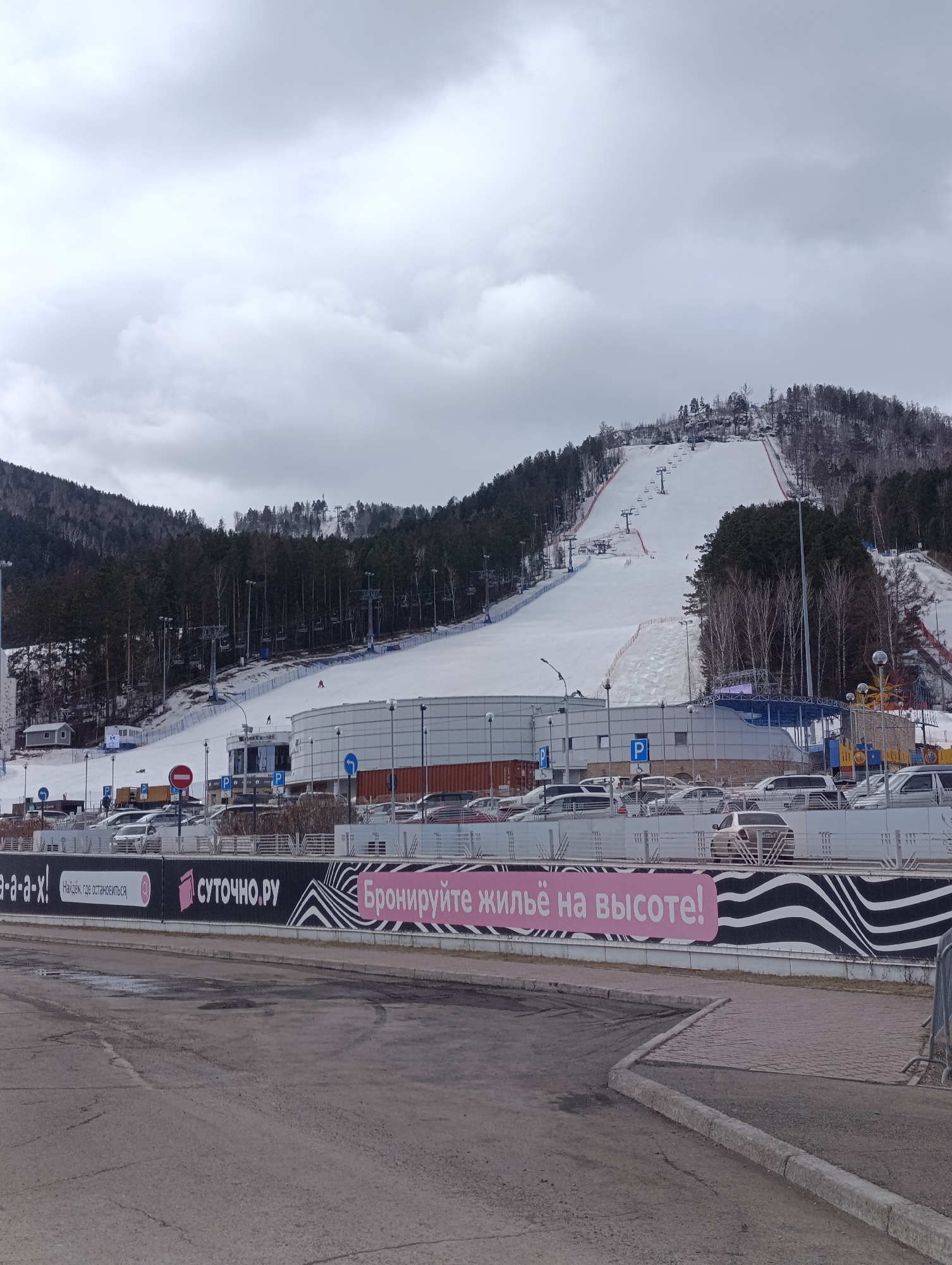 The image shows the Bobrovy Log ski resort in Krasnoyarsk, Russia on an overcast day. A wide ski slope covered in snow extends up a tree-covered mountain, with ski lifts visible. At the base of the slope is a multi-story building, a parking lot filled with cars, and a road in the foreground. A banner with the text "СУТОЧНО РУ Бронируйте жильё на высоте!" (Rent accommodation at a height!) is visible along the bottom edge of the image.