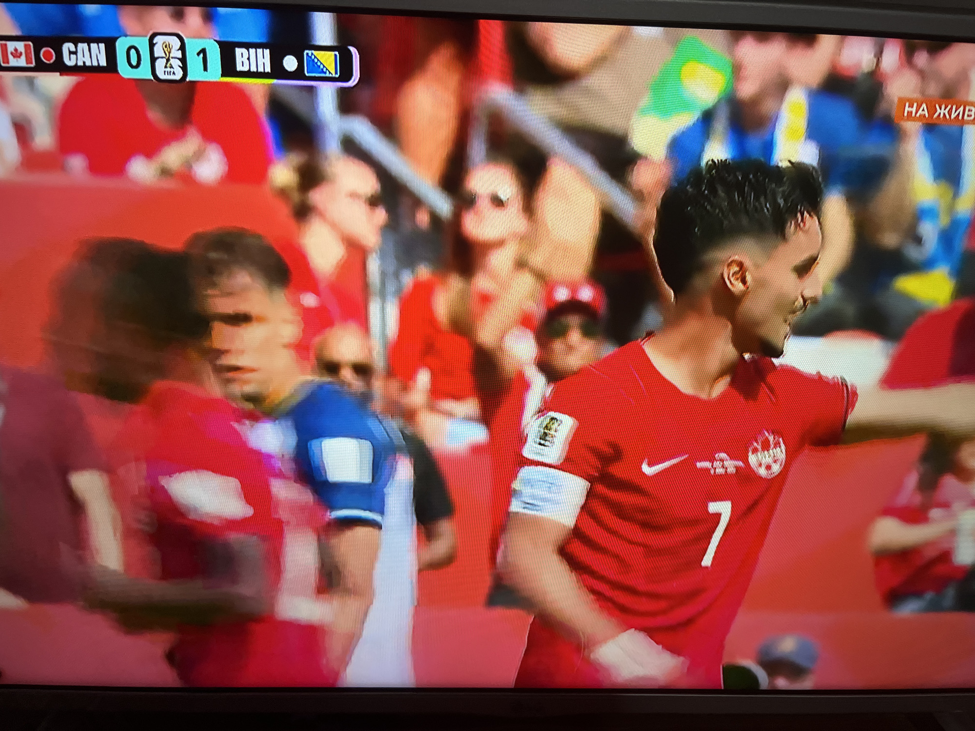 In a digital television screen display, a soccer match scoreboard shows "CAN 0 1 BIH" with national flags of Canada and Bosnia and Herzegovina. The camera focuses on a male soccer player in a red jersey with the number 7 on it, playing in what appears to be a stadium setting. The background shows blurred images of spectators and stadium elements, indicating the match is in progress. The player is looking to his right and appears to be in mid-action. The overall scene is vibrant and dynamic, capturing a moment from a live sporting event.