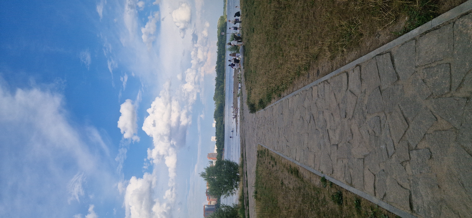 This image depicts a paved walkway made of irregular stone slabs leading towards a body of water, likely the Ob River, on a partly cloudy day. On the far bank, a dense line of trees separates the river from a cityscape featuring several tall, multi-story buildings. Along the near bank, a small group of people are gathered, some appearing to be standing by the water's edge. The foreground is dominated by the pathway and dry, sparse grass on either side, suggesting a warm season. The sky is a vibrant blue with scattered white cumulus clouds. The scene captures a public space within the city of Novosibirsk, Russia, likely a park or waterfront area.