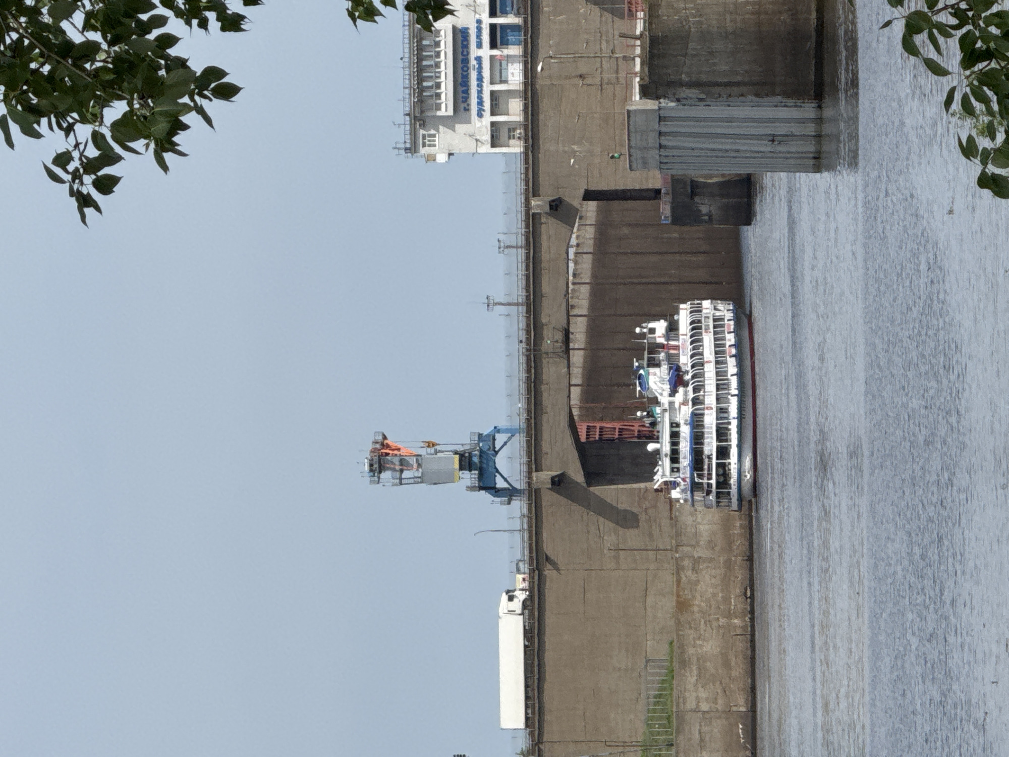 The image shows a river lock, with a passenger ship inside the lock chamber. The river lock appears to be in the city of Chaikovsky, Russia, based on the inscription on the building in the background, which reads "Чайковский" (Chaikovsky). The lock is made of concrete and steel, and the water level in the lock is at or near the river level. There is a crane next to the river lock. The passenger ship is white with multiple decks. There are trees visible in the foreground. The sky is a clear blue, indicating sunny weather.