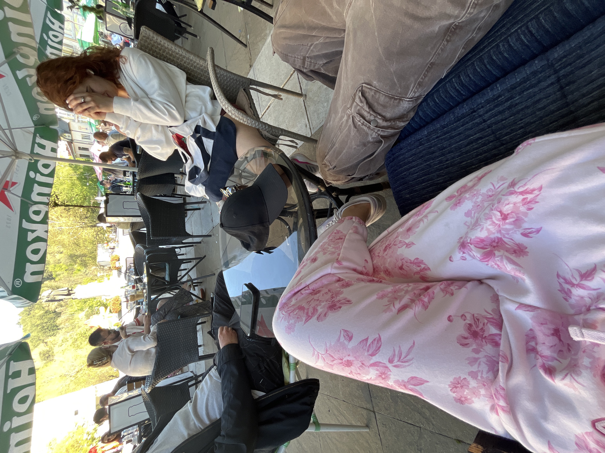 The image shows an outdoor scene at a cafe, likely in Blagoevgrad, Bulgaria based on the cityscape and surrounding architecture. There are multiple people seated around tables with a glass top. A woman with red hair, wearing a white sweater, has her hands covering her face. Others are dressed in casual wear, including cargo pants, jeans, and a pink floral-patterned outfit. A black baseball cap sits on the table. Some people at other tables in the cafe are partially visible, seated on black, woven chairs. A green umbrella, branded with "Heineken," is visible in the background and to the left, presumably providing shade. A variety of objects are on the tables, likely drinks.