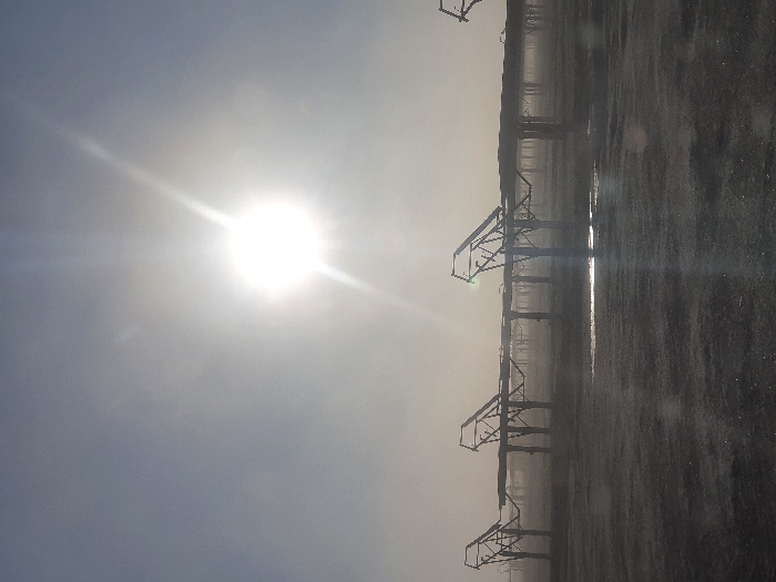 A bright sun shines in a hazy sky, partially obscured by clouds. Silhouetted against the glare are structures resembling irrigation systems or industrial equipment. They appear in a linear formation along a gray, reflective surface, possibly wet ground or standing water, creating mirroring effects. The scene is dominated by the sun's luminescence, resulting in low contrast and obscuring details in the foreground. The overall impression is desolate and slightly ethereal.