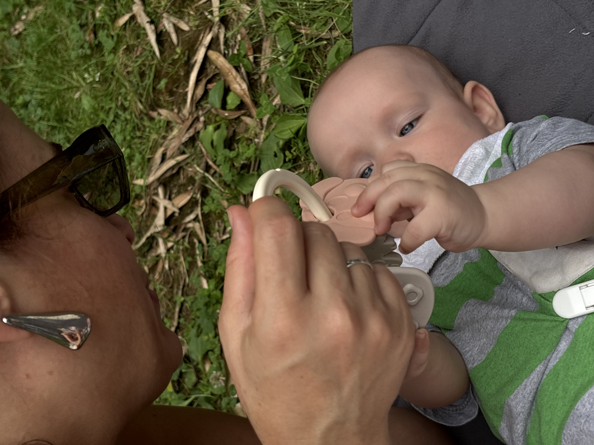 A close-up, eye-level shot shows a woman leaning over a baby outdoors. The woman, partially visible from the shoulders up, wears glasses with dark frames and a large, metallic, teardrop-shaped earring in her left ear. Her right hand holds a pink teether toy with a white ring, offering it to the baby. The baby, lying on its back on a dark gray blanket, wears a gray and green striped shirt. The baby's left hand reaches up towards the toy, and its right hand is near its mouth. The background consists of green grass, dried leaves, and scattered foliage, suggesting a park or garden setting. The scene does not provide any specific visual cues to indicate its location within the city of Sochi, Russia.