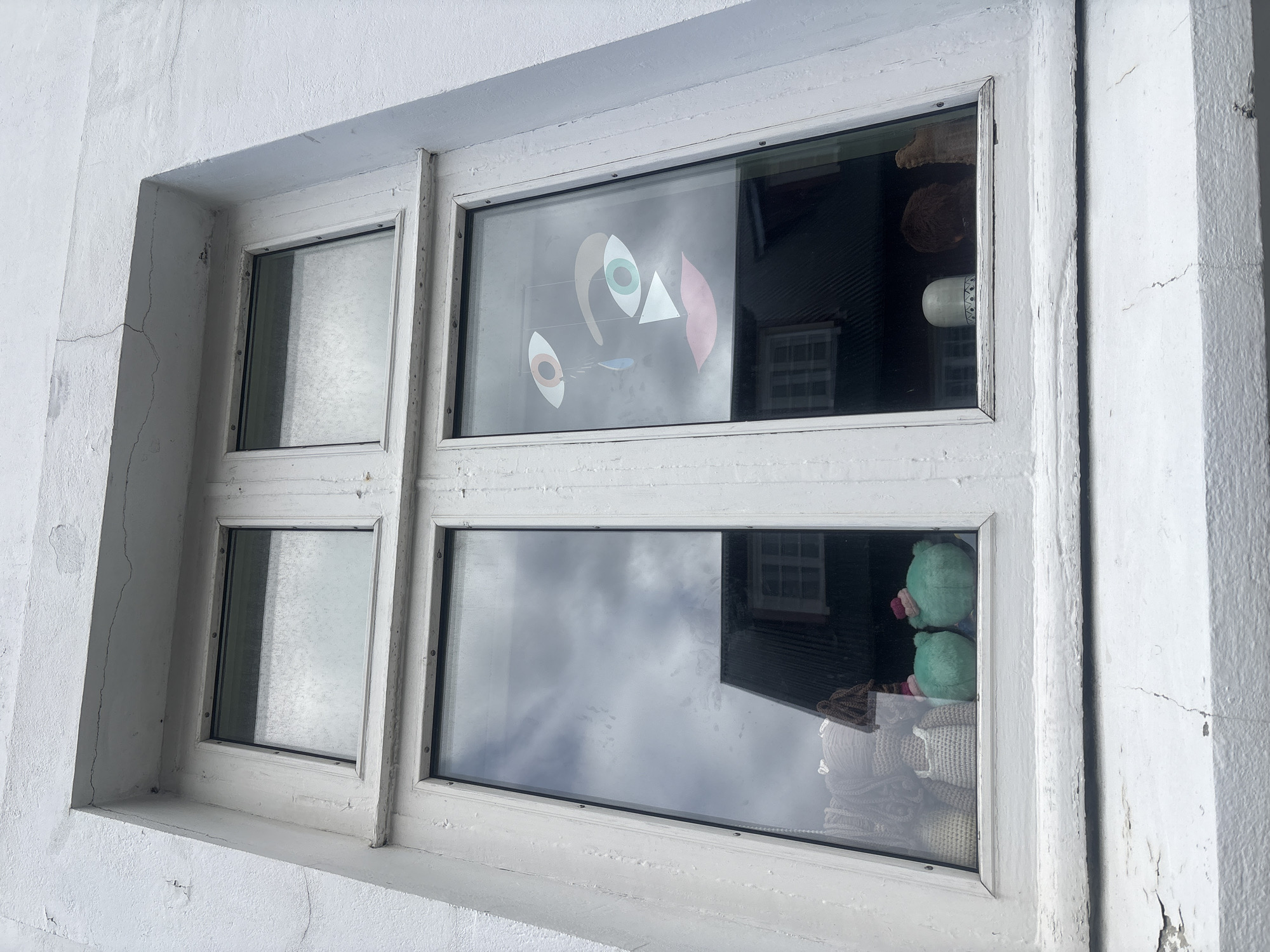 The image shows a closed, white-framed window on the exterior of a building with a textured white facade. The window is divided into four panes, with two larger panes on the bottom and two smaller panes on top. The panes are reflective, showing a cloudy sky and the dark, sloped roof of a building across the street.
In the right-hand pane, a colorful art installation depicting a stylized face is visible. It features large, geometric eyes, one blue teardrop, and pink lips. In the bottom left pane, several crocheted figures are lined up, including what appear to be two small, round, green plush toys with red hats, and several beige figures. In the bottom right pane, a white ceramic cylindrical object with blue patterns is visible, along with a fuzzy brown object and a piece of driftwood.
The building's facade exhibits signs of age, with visible cracks and peeling paint, particularly along the bottom edge of the window frame. The overall impression is of a weathered urban environment, likely a residential or commercial building.