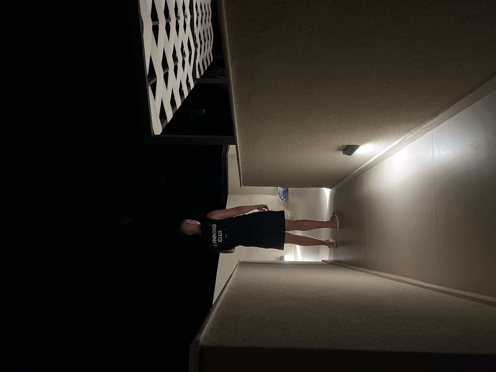 A person stands with their back to the camera, walking down a dimly lit, outdoor walkway at night. They are wearing a black sleeveless shirt with "RUNNING CLUB" in white lettering on the back, black shorts, and white flip-flops. The walkway is bordered by textured walls on both sides, with upward-facing lights illuminating the path. Above, a white lattice-like structure with dark supports is visible against the black night sky. The overall scene suggests an outdoor architectural space in Sithonia Municipal Unit, Greece.
