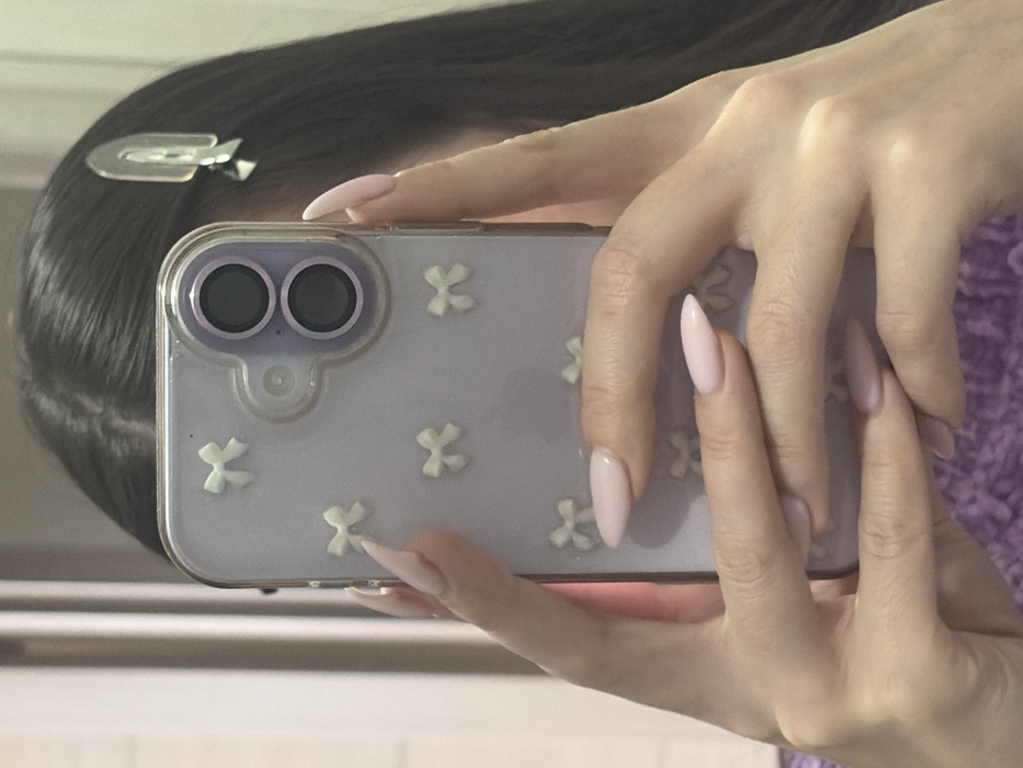 The scene is located in Novosaratovka, Russia. The image displays a close-up, reflected view of an individual, likely female, from the upper torso to just above the head. The subject's long, dark hair is parted down the center, with a clear or light-colored, arched hair clip positioned on the right side of the head. Both hands are holding a smartphone vertically, with fingers displaying long, pointed nails painted in a light pink or nude shade. The smartphone is enclosed in a translucent purple case featuring multiple small, white, three-dimensional bow embellishments. The rear of the smartphone shows a camera module with two vertically stacked dark lenses. A textured purple garment, presumably worn by the individual, is visible in the lower portion of the image. The background is out of focus and indistinct, consistent with a reflection in an indoor setting.
