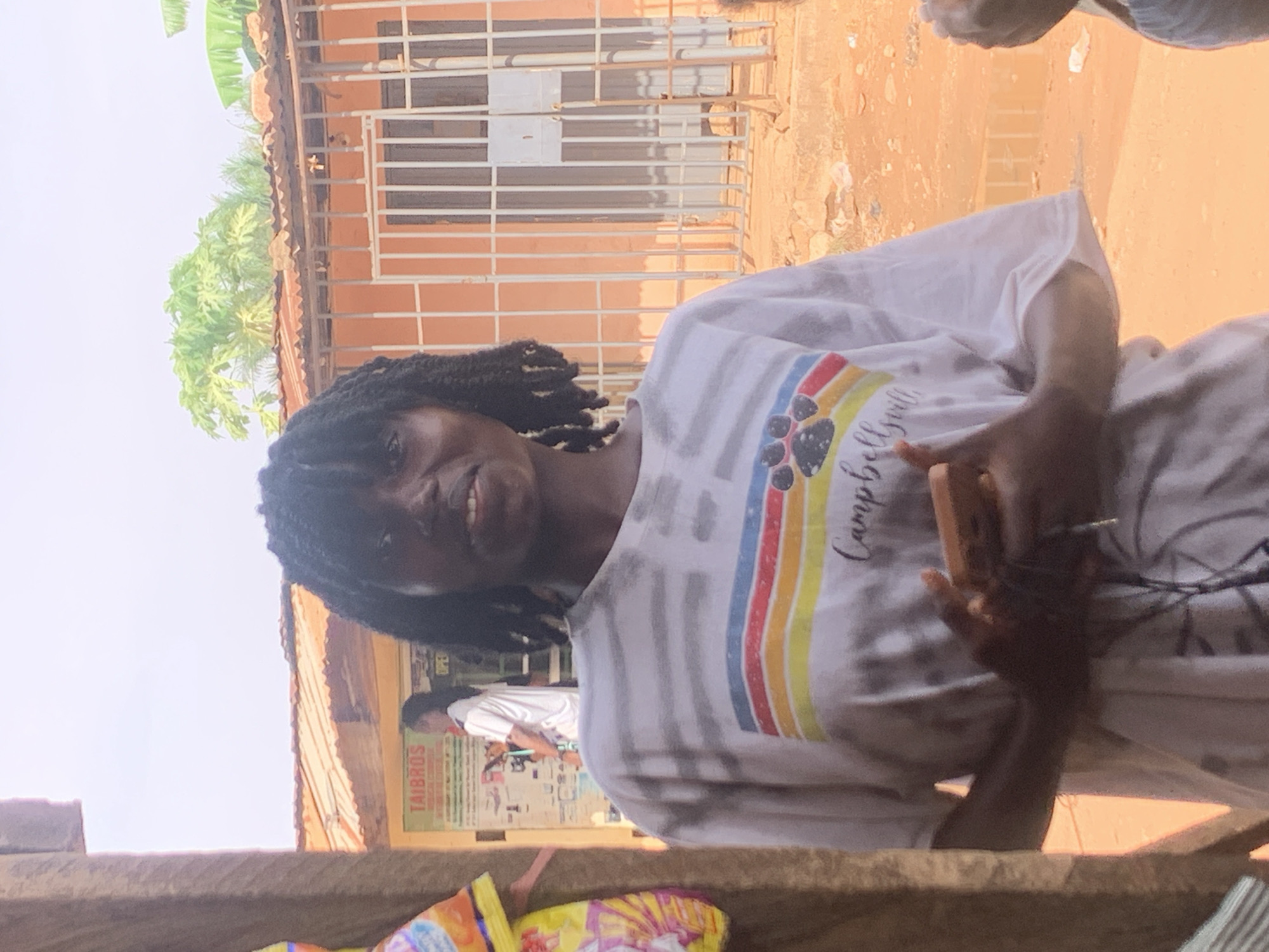 A young person with dark skin and dreadlocks is captured in a medium close-up, smiling towards the camera. They are wearing a light-colored, possibly white or light grey, short-sleeved t-shirt featuring a prominent horizontal rainbow stripe design. The stripe includes colors such as red, orange, yellow, green, light blue, and dark blue. Within the rainbow stripe, a dark paw print is visible, and the text "Campbell Girls" is written in a cursive font. The person is holding a smartphone in their left hand, which appears to be dark-colored.
The background suggests an outdoor, possibly semi-urban or rural setting, consistent with Sabon Tasha, Nigeria. Behind the subject, a building with reddish-brown walls is visible. Part of the building features a window with white metal bars. Above the building, a corrugated iron roof is discernable, and lush green foliage, resembling banana and papaya trees, rises into the frame against a bright, clear sky, indicating daytime. Another person, dressed in a white t-shirt, is visible in the distance to the left, standing near a sign that reads "TAIBROS" in large green letters, with other smaller, unreadable text below it. The ground or a low wall in the foreground is a rough, reddish-brown texture. The overall mood is casual and bright, with the subject displaying a pleasant expression.