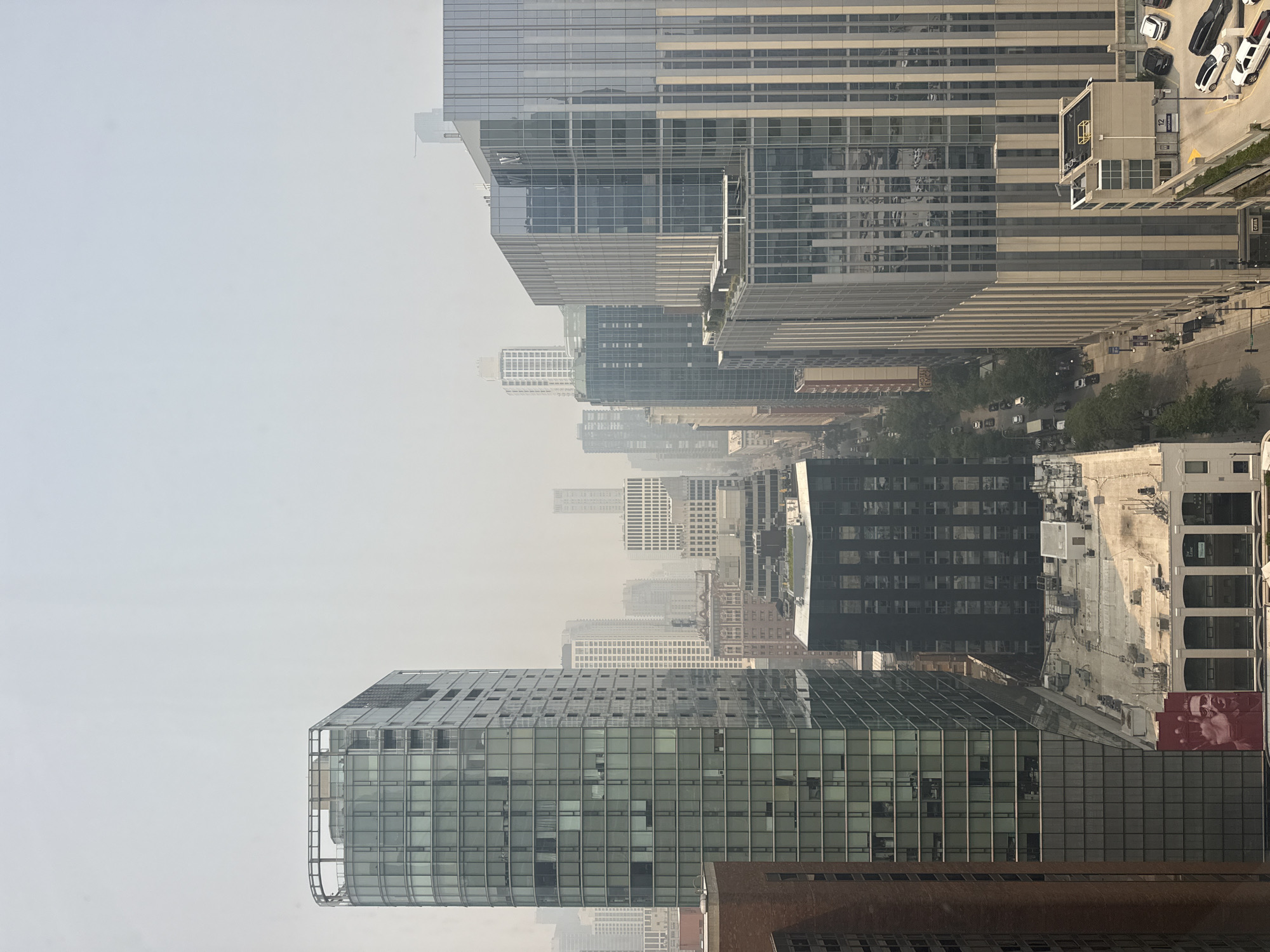 The image depicts a hazy urban landscape in Chicago, Illinois, United States of America. The view is from an elevated perspective, looking down onto a street lined with tall buildings of varying architectural styles.
In the foreground on the left, a modern, glass-faced skyscraper dominates the view. Its facade is a mosaic of rectangular windows and vertical elements. Adjacent to it, to its right, is another, even larger skyscraper with a more uniform, vertical striped pattern of glass and metal. This building reflects the sky and surrounding structures. Further to the right, a parking garage structure with visible cars is present.
The street level shows a typical urban scene with cars, trees lining the sidewalks, and pedestrians. Some of the buildings have visible signage, indicating businesses or residential units. The overall atmosphere is muted, likely due to atmospheric haze or smoke, which obscures distant details and gives the sky a milky appearance.
On the facade of one of the buildings on the left, a large red mural featuring two faces and hands is visible. The mural appears to be a prominent piece of street art within the cityscape. The presence of a mural suggests an artistic and cultural element within this part of the city.
The buildings exhibit a mix of contemporary and older designs, with flat roofs, some featuring rooftop amenities like greenery and lounge areas. The density of the urban environment is evident in the close proximity of the buildings and the multi-lane street below. The overall impression is that of a busy metropolitan area experienced on a day with limited visibility.
