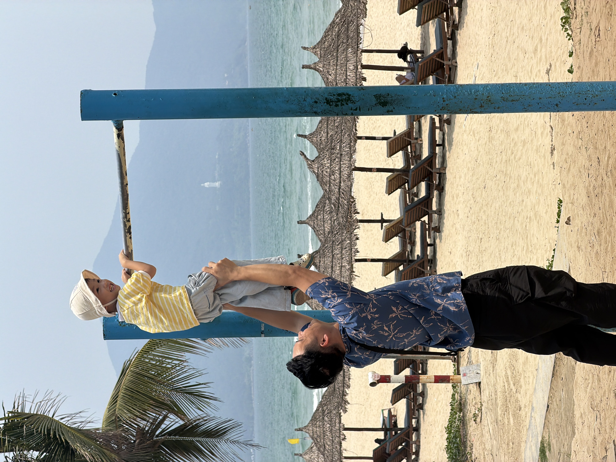 A medium shot features a man holding a small child hanging from a blue metal bar, with a beach scene as the background. The man has black hair and is wearing a blue shirt with a leafy pattern and black pants. The child is wearing a yellow striped shirt, grey shorts, and a light-colored hat. The man is lifting the child so they can hang from the bar. The background includes sandy beach, rows of beach umbrellas, and lounge chairs, along with the ocean and distant mountains. The location is likely a beach in Đà Nẵng, Vietnam.