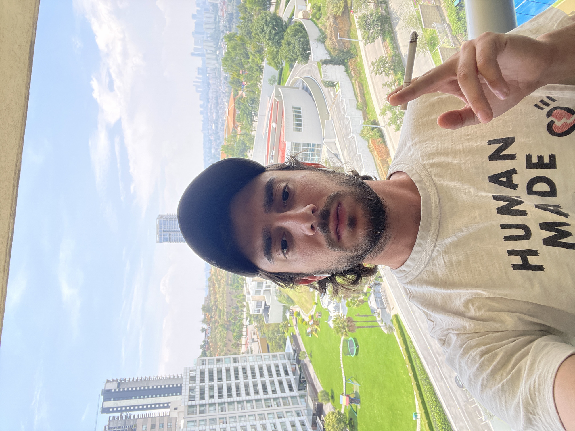 A man with a beard and a black baseball cap is posing for a selfie while holding a lit cigarette in his fingers. He is wearing a white t-shirt with the text "HUMAN MADE" in black. Behind him is a view of Huixquilucan de Degollado, Mexico, which includes buildings, trees, a green lawn with playground equipment, roads, and a cloudy sky.