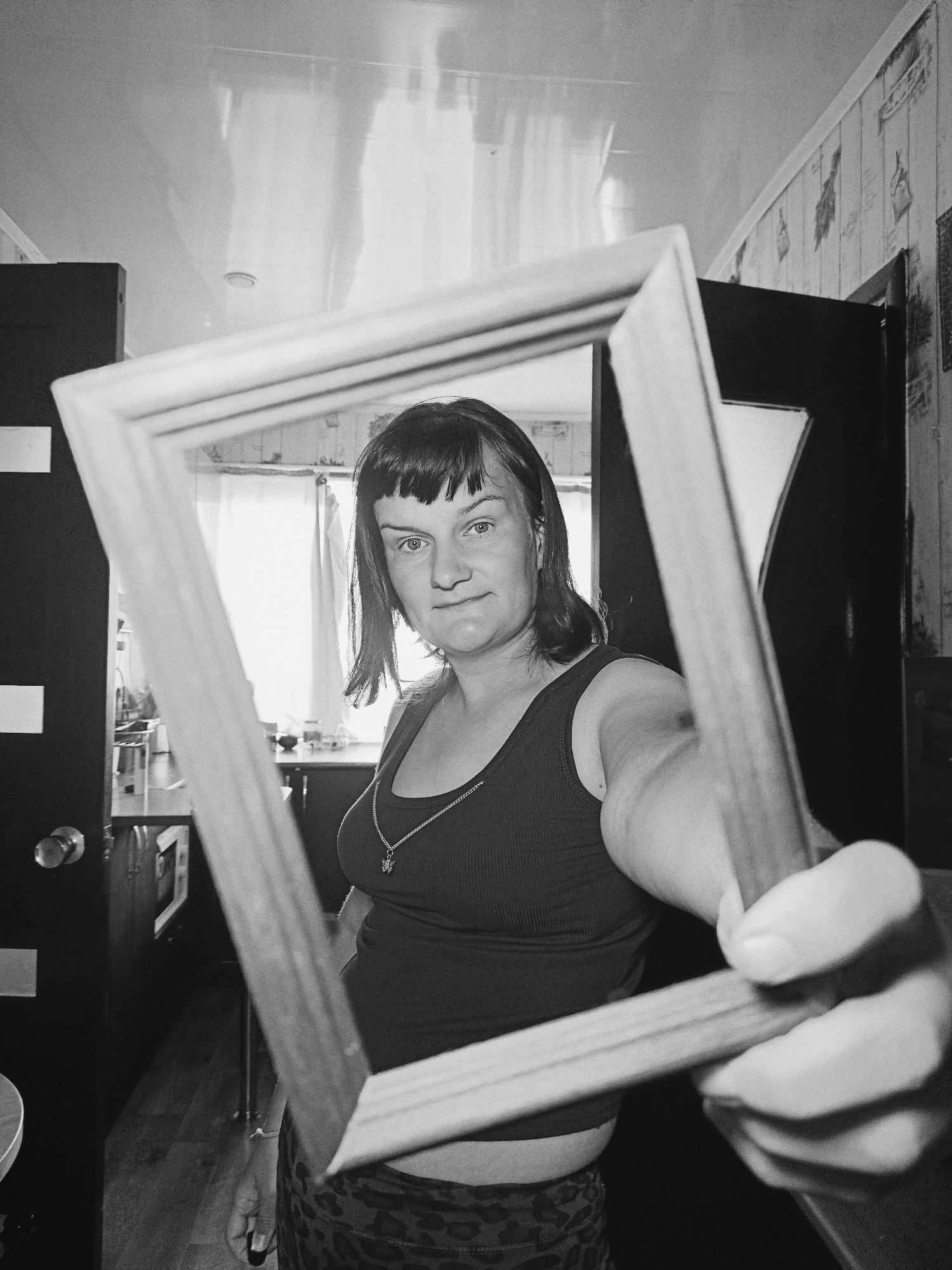 A monochrome, low-angle shot depicts a woman indoors, holding a square wooden frame in front of her. She gazes at the camera through the frame. The woman has dark hair, blunt bangs, and is wearing a tank top and a necklace. Her patterned pants are visible. The frame is held with her right hand, obscuring the lower portion of her body. In the background, a doorway leads to another room with a window and furniture. Another door is visible to the left. The location and city are Unknown, Unknown.