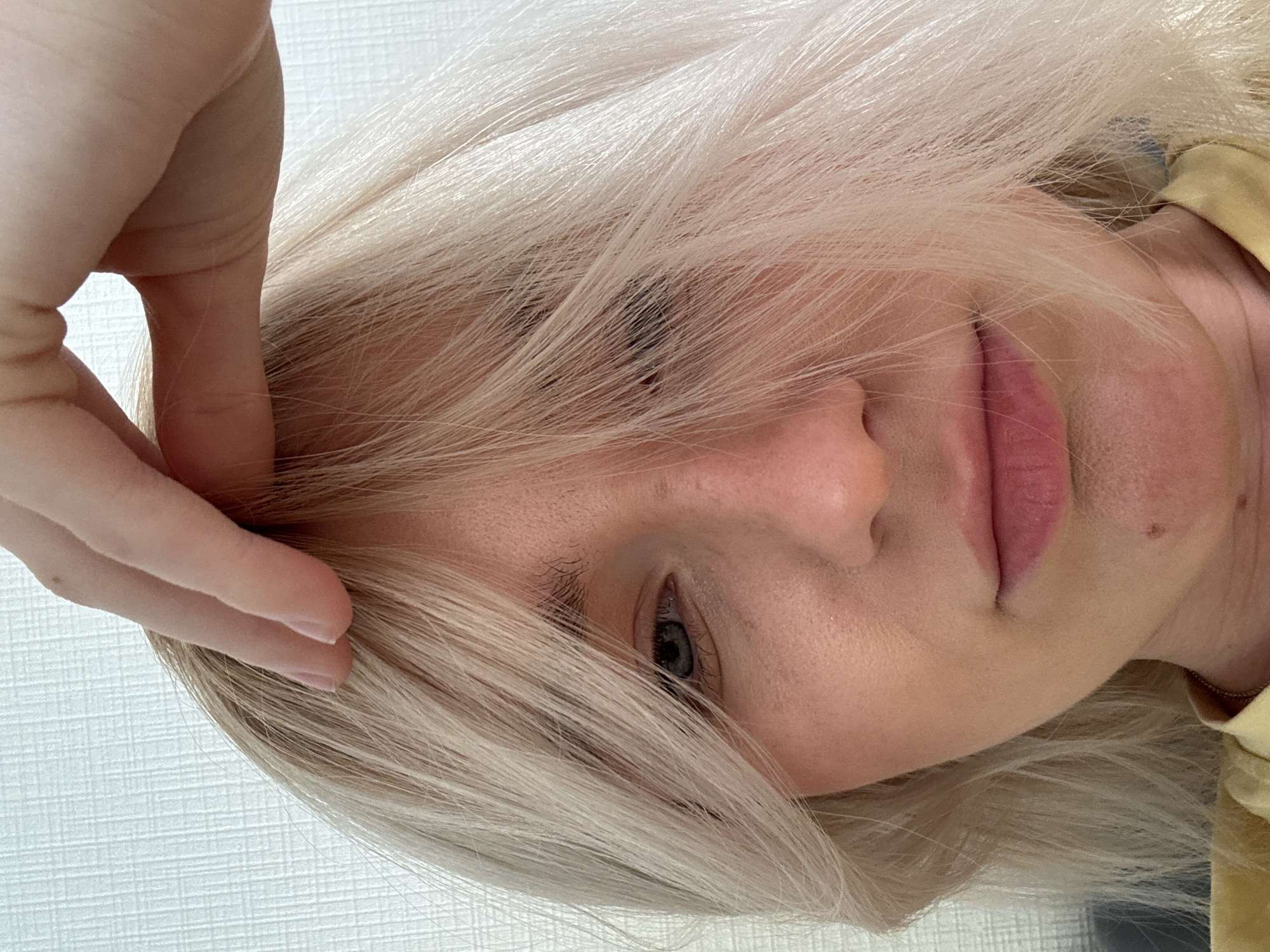 A close-up, slightly rotated image depicts a person with fair skin and blonde hair partially obscuring their face. A hand is positioned near their forehead, seemingly touching or adjusting their hair. The person's left eye is visible and appears to be blue or gray. Their lips are pink. A small mole is visible near their mouth. The background appears to be a plain, light-colored wall. There is no evidence indicating this image was taken in the city of Vsevolozhsk, Russia.
