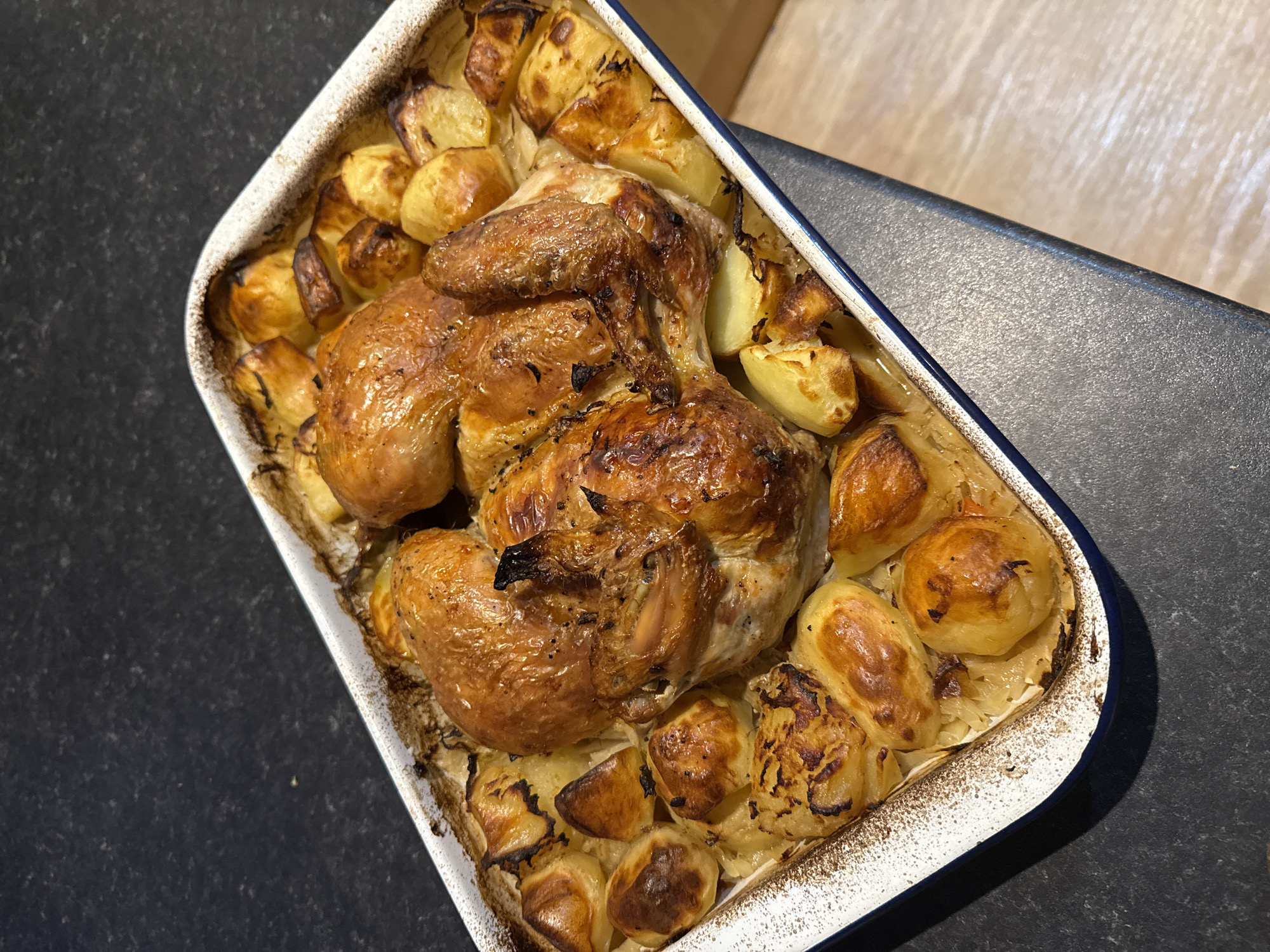 The main subjects of the image are two whole roasted chickens and numerous roasted potatoes, presented in a white rectangular baking dish with a blue rim. The chickens have a rich golden-brown, crispy-looking skin, suggesting they are perfectly cooked. They are positioned centrally within the dish, surrounded by large, irregularly shaped roasted potato pieces, which also exhibit an appetizing golden-brown color with some darker, caramelized spots. Beneath the poultry and potatoes, a layer of finely chopped or sliced cooked vegetables, possibly onions, is visible, along with savory pan juices that have accumulated at the bottom of the dish.
The baking dish itself is white, made of ceramic or enamelware, and features a noticeable blue band along its top edge. Its sides bear marks of use, with baked-on brown residue from the roasting process, indicating it has just come out of an oven. The dish is placed on a dark, speckled countertop, which occupies the lower left and foreground of the scene. A lighter, horizontally-grained wooden surface is visible in the background on the right, suggesting the setting is a domestic kitchen or dining area.
The overall scene depicts a hearty, home-cooked meal, likely a Sunday roast or a family dinner, prepared and ready for serving. The time of day is implied to be mealtime, either lunch or dinner. The food appears inviting and well-prepared, evoking a sense of warmth and comfort. No visible text is present in the image. The location context provided is Great Yarmouth, United Kingdom, aligning with the commonality of such a meal in a UK household.