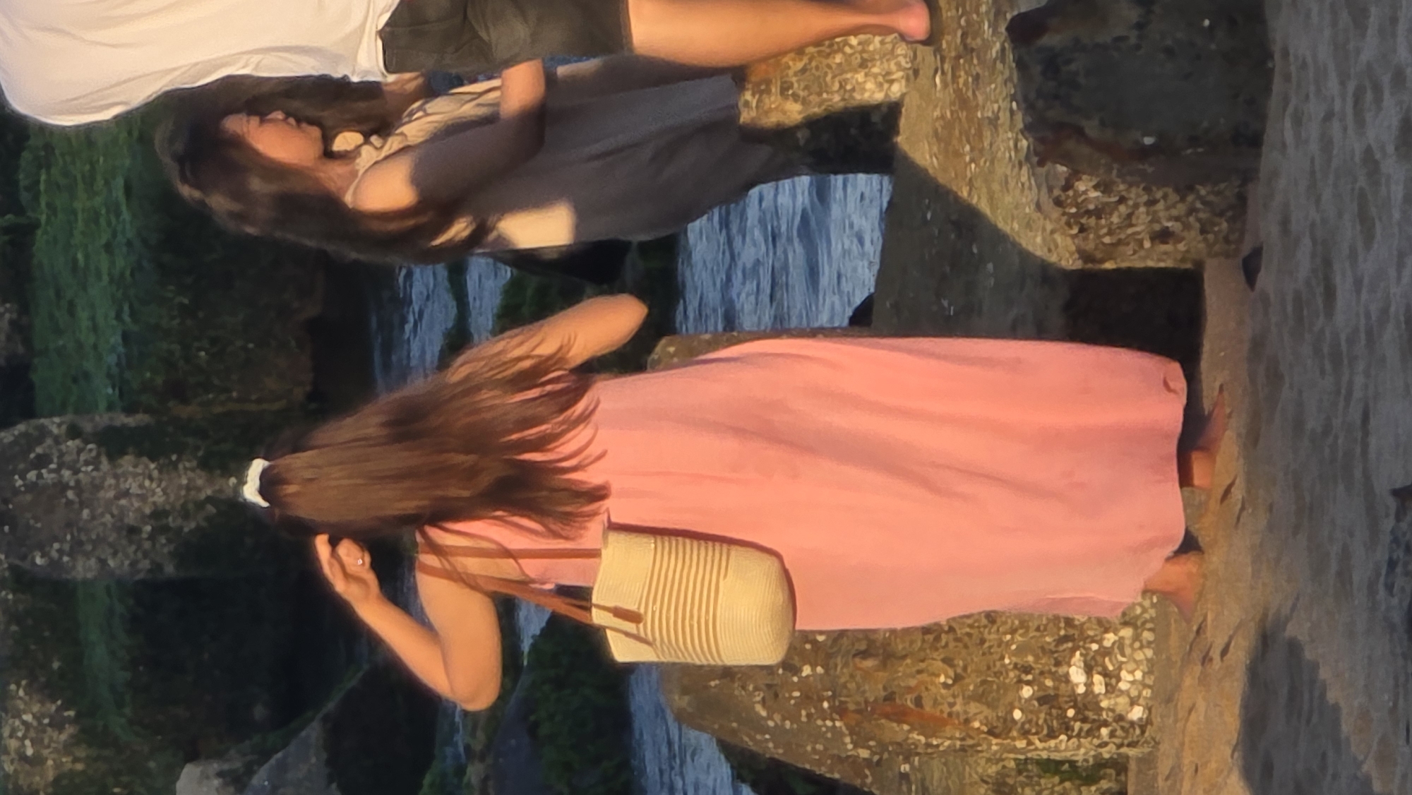 A long-haired woman in a pink dress and a striped bag stands on a rocky outcrop overlooking a body of water. A second woman, wearing a tan dress, stands with a man on similar rocks nearby. The location appears to be in Nha Trang Ward, Vietnam.