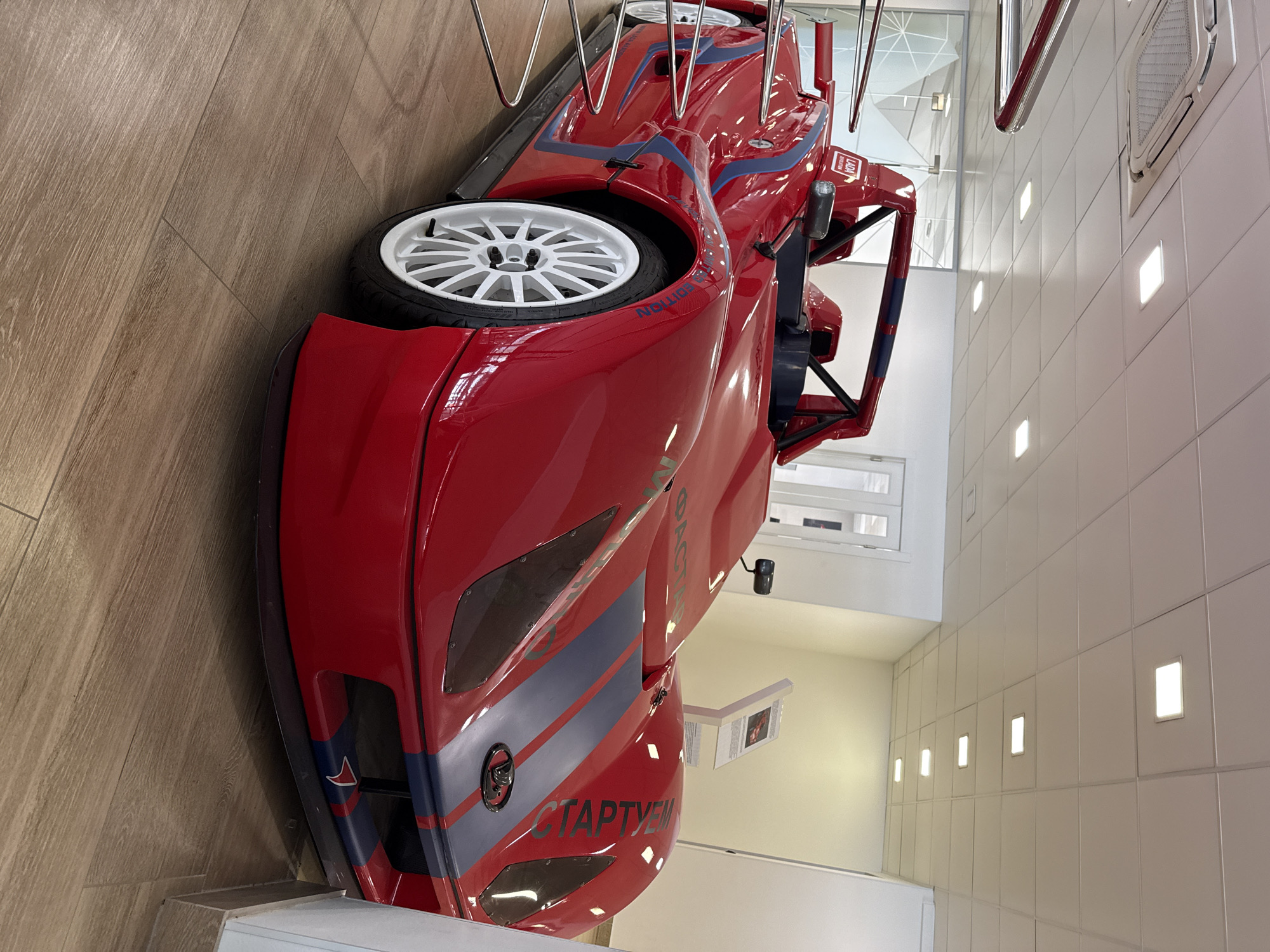 The image shows a red race car on display in a room with light wood flooring and white tile walls. The car has white rims, blue and gray stripes, and the word "СТАРТУЕМ" in Cyrillic script on the side. The car is surrounded by a metal railing. The room is well-lit with recessed lighting in the ceiling.