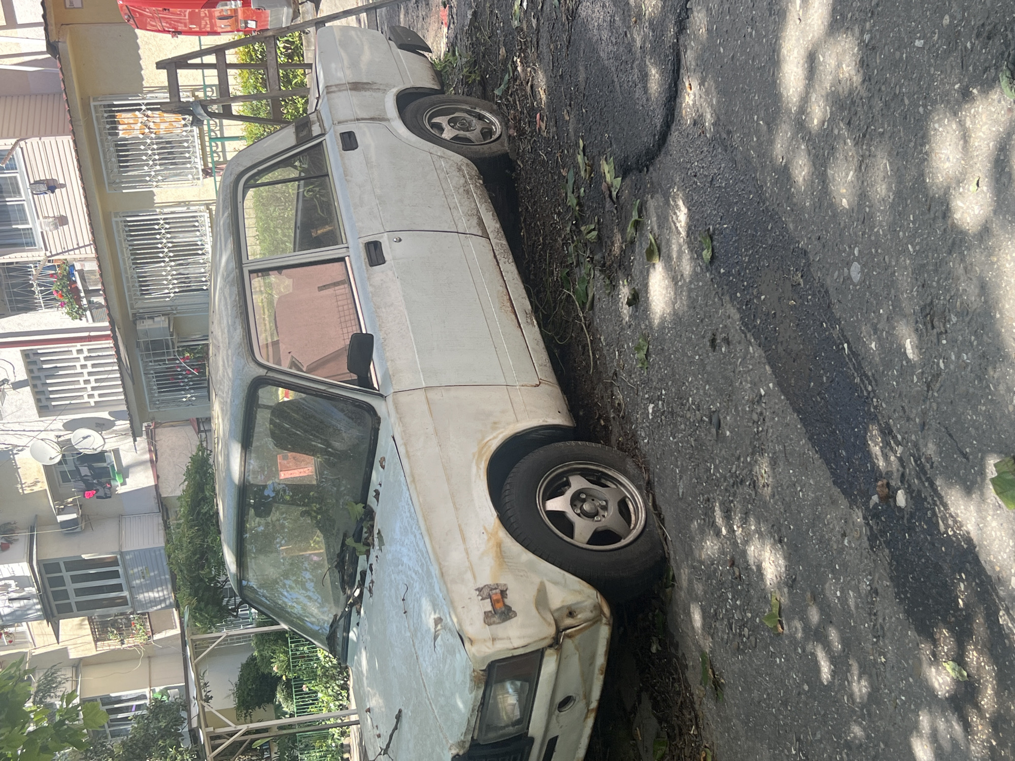 The image, captured in Sochi, Russia, displays an outdoor scene featuring a white, heavily weathered sedan, likely an older Soviet-era model such as a Lada. The car, positioned partially on an unpaved patch next to an asphalt road, exhibits extensive rust staining along its body panels, particularly on the doors, fenders, and roof. The front right fender is visibly damaged, with bent metal and a broken or missing turn signal lens. It features aftermarket five-spoke alloy wheels. Debris and leaves are scattered across its hood and roof, and small plants grow near its base, indicating potential disuse or abandonment.
In the background, several multi-story residential buildings are visible, characterized by balconies, some adorned with decorative window grilles or bars. Satellite dishes and air conditioning units are mounted on their facades. One building is a pale yellow, while another features light-colored horizontal siding. A small portion of a red vehicle can be seen in the upper right corner, partially obscured by a wooden structure.
The scene is well-lit by direct sunlight, casting dappled shadows from unseen trees onto the asphalt road and surrounding areas. Fallen leaves are scattered on the ground. The setting appears to be a quiet, possibly residential or alleyway environment during daytime. No people or discernible text are present in the image.