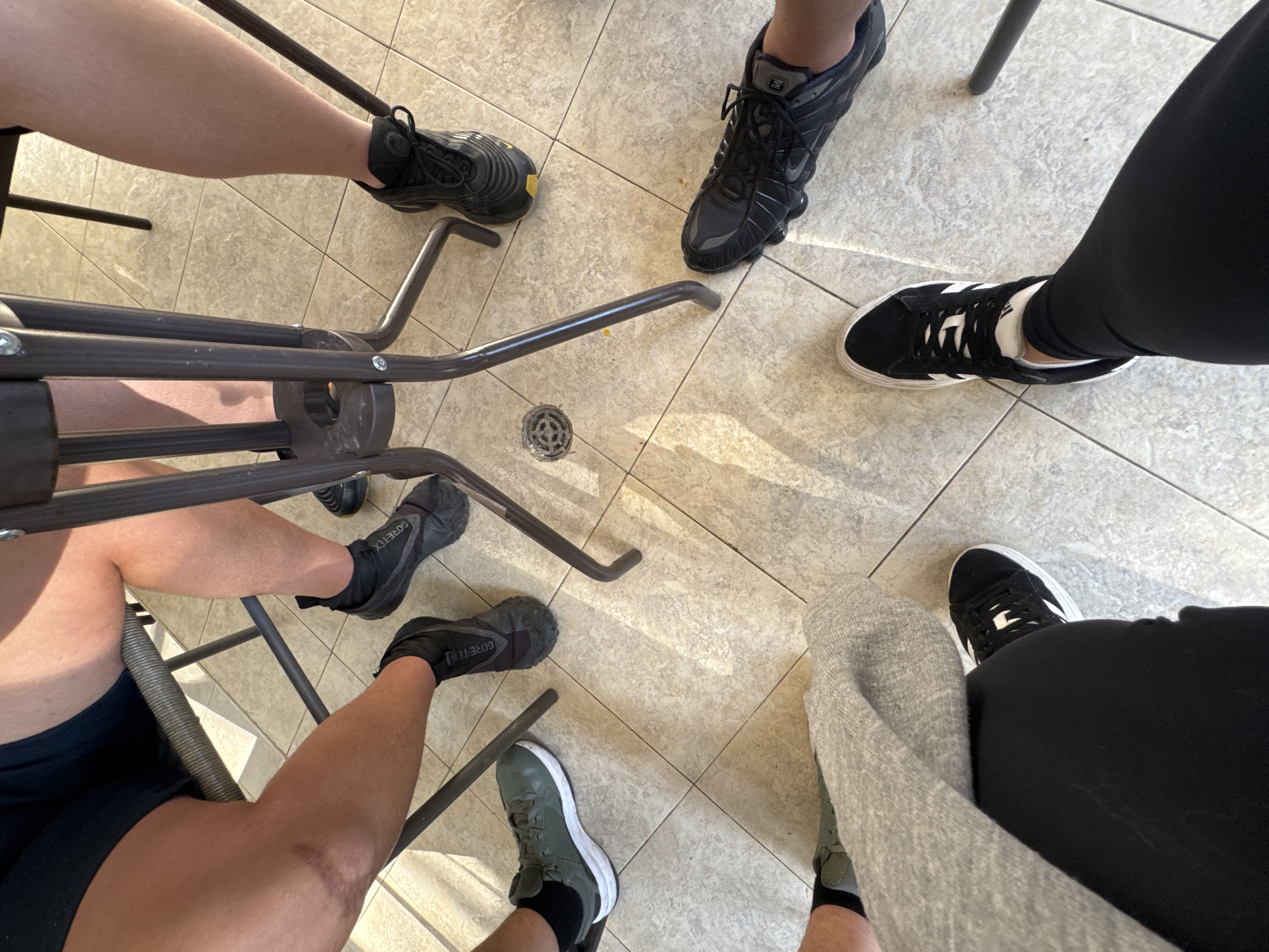 The image shows a group of people sitting around a table, with their feet and lower legs visible. The location appears to be indoors on a tiled floor. Several pairs of sneakers are visible, including black and gray athletic shoes and green sneakers. The legs are clad in shorts, leggings, and a gray sweater. The table has a metal base. A drain is visible in the floor.
