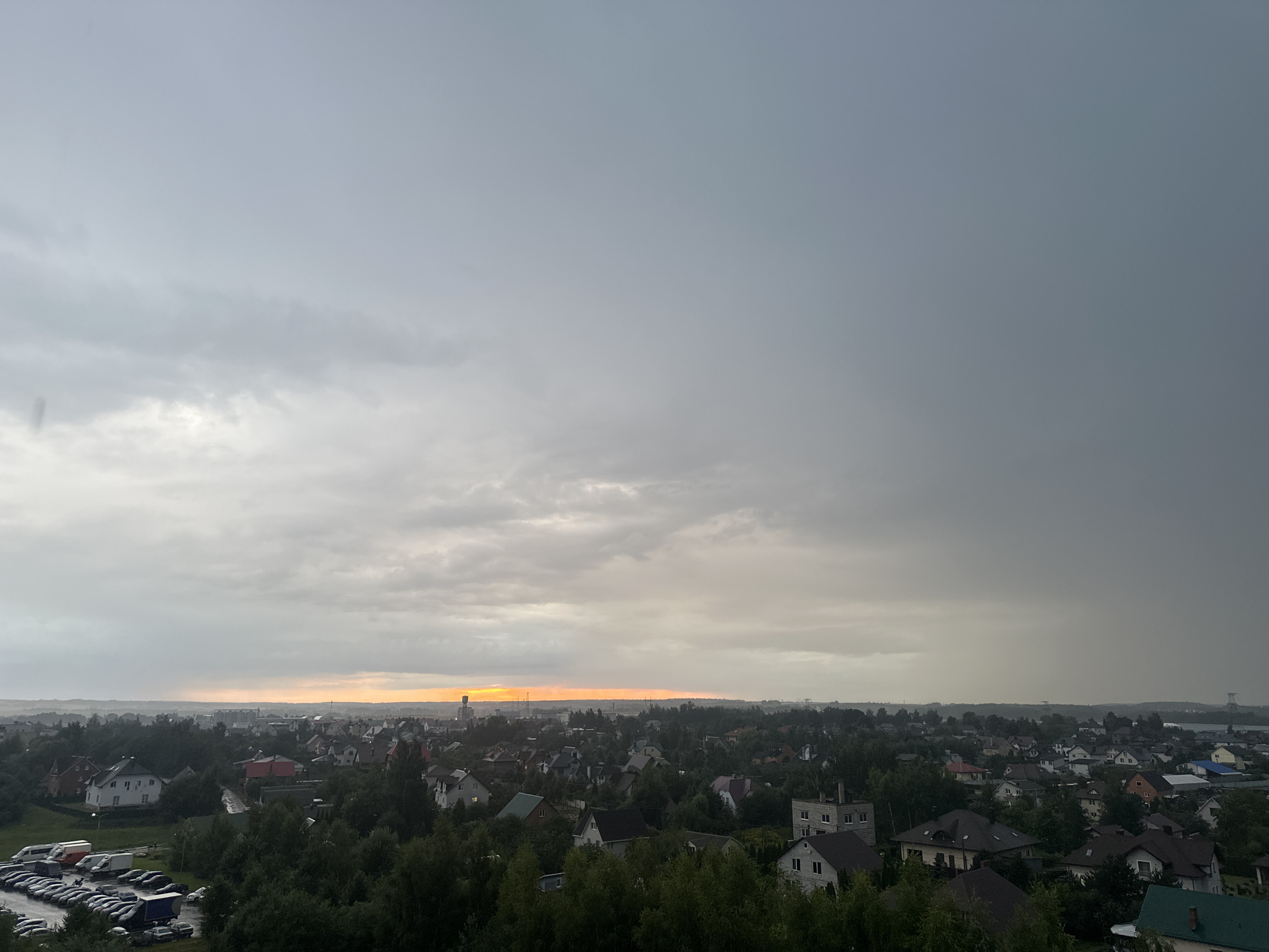 This is an aerial shot of a city during a cloudy day, possibly at sunset or sunrise, with a light orange glow visible on the horizon. The city, identified as being in the vicinity of Шчомысліцкі сельскі Савет, Belarus, is densely packed with houses of various sizes and architectural styles, many with pitched roofs. Lush green trees are interspersed throughout the residential areas, some reaching up to the rooftops. In the foreground, on the left side of the image, there is a large parking lot filled with numerous vehicles, including vans and cars. The sky is predominantly covered with heavy, gray clouds, with a thin band of lighter, warmer colors near the horizon. A tall, slender tower-like structure is visible in the distance on the right side of the frame. The overall atmosphere suggests a somewhat overcast and possibly humid environment.