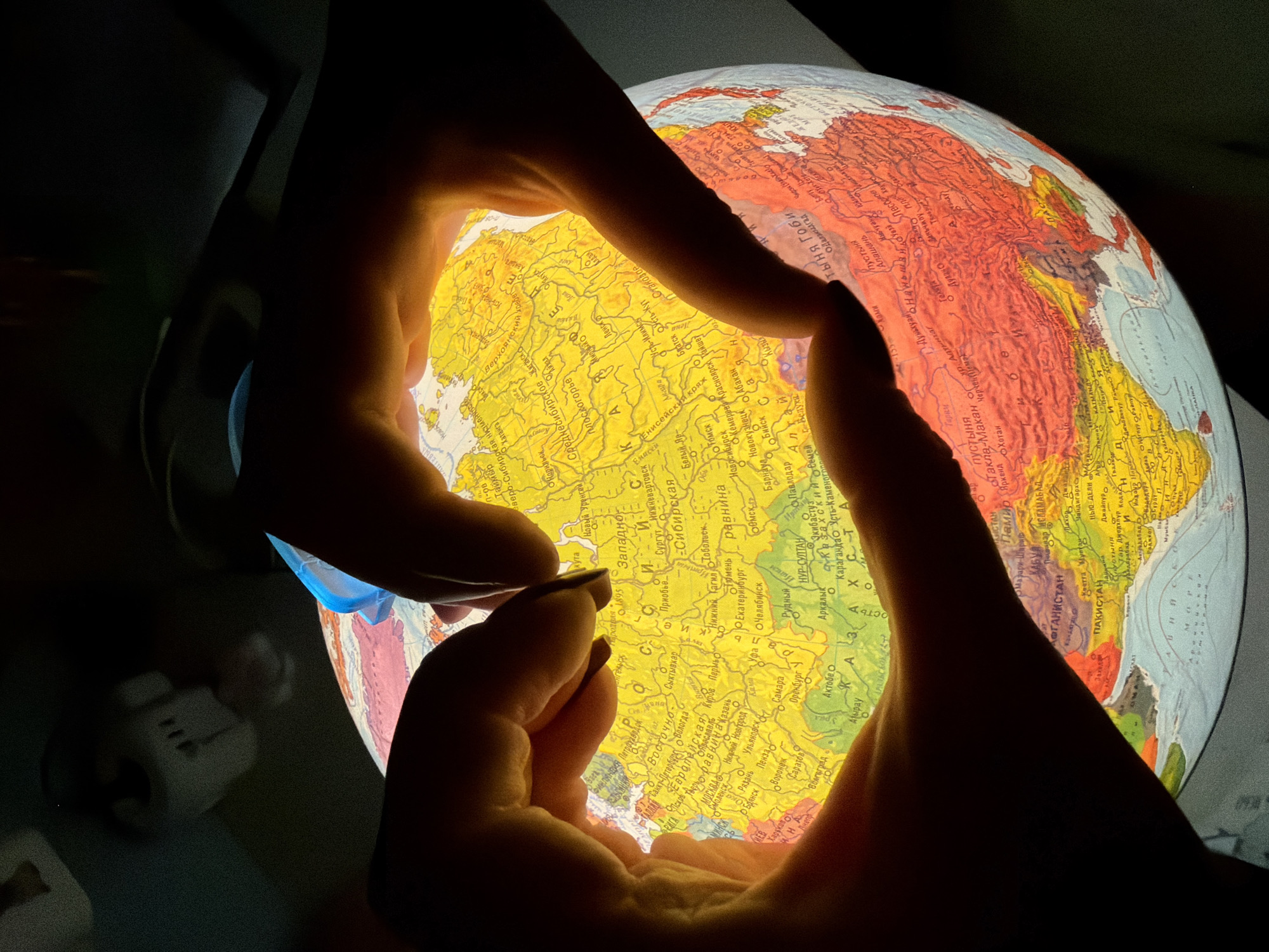 In this close-up shot, two hands are illuminated by the internal light of a globe, creating a heart shape as they hold it. The globe displays a map of Asia, with Russia prominently featured. The hands are silhouetted against the bright light of the globe, obscuring the face and body of the person holding it. The overall scene is dimly lit, with the glowing globe serving as the primary light source. The map on the globe shows various cities and geographical features in Russia and surrounding countries, with text in Cyrillic. Some of the visible text includes "Россия" (Russia), "Сибирь" (Siberia), and names of cities like "Москва" (Moscow) and "Санкт-Петербург" (Saint Petersburg). The image evokes a sense of wonder and connection to the world.