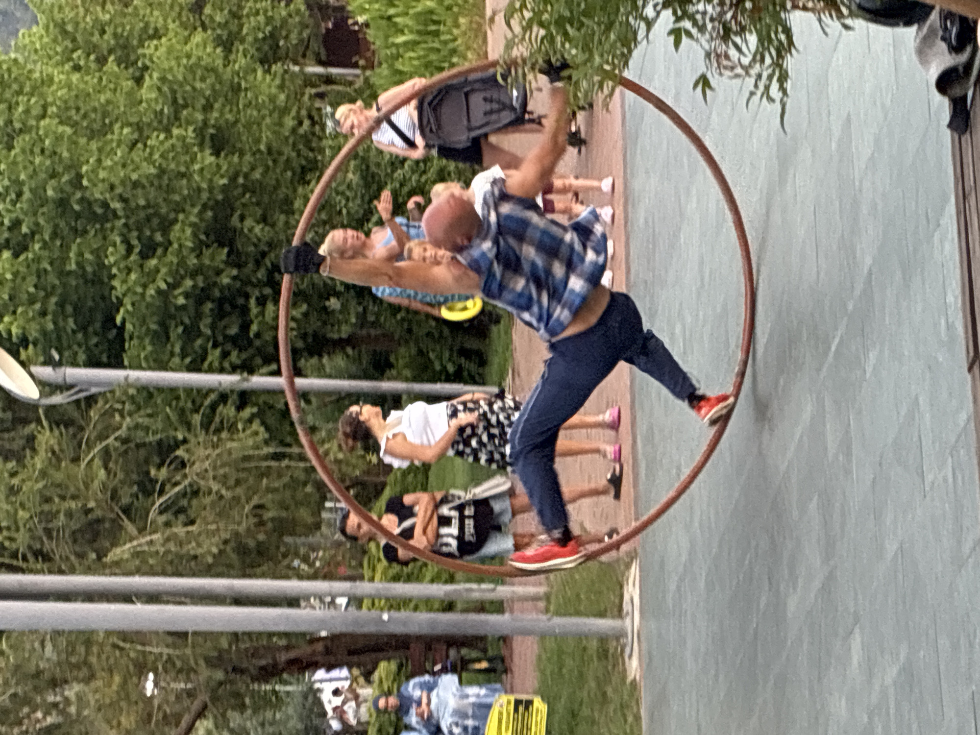 An acrobatic performer is captured mid-performance in a large circular hoop, performing a dynamic maneuver. The performer, wearing a blue and white plaid shirt and dark pants with red shoes, is positioned inside the hoop, with one arm extended upward and the body angled as if spinning. The hoop, a reddish-brown color, is suspended and appears to be part of the act.
In the background, several individuals are watching the performance. To the left, a woman in a blue patterned dress sits on a bench. Behind the performer, a group of women, some with children, observe the spectacle. The environment suggests an outdoor public space, possibly a park or promenade. Tall, slender vertical poles are visible, with greenery and trees forming a backdrop. The ground is paved with grey rectangular tiles. The scene takes place in Alanya, Turkey, likely a public area designed for leisure and entertainment, given the presence of spectators and the acrobatic performance.