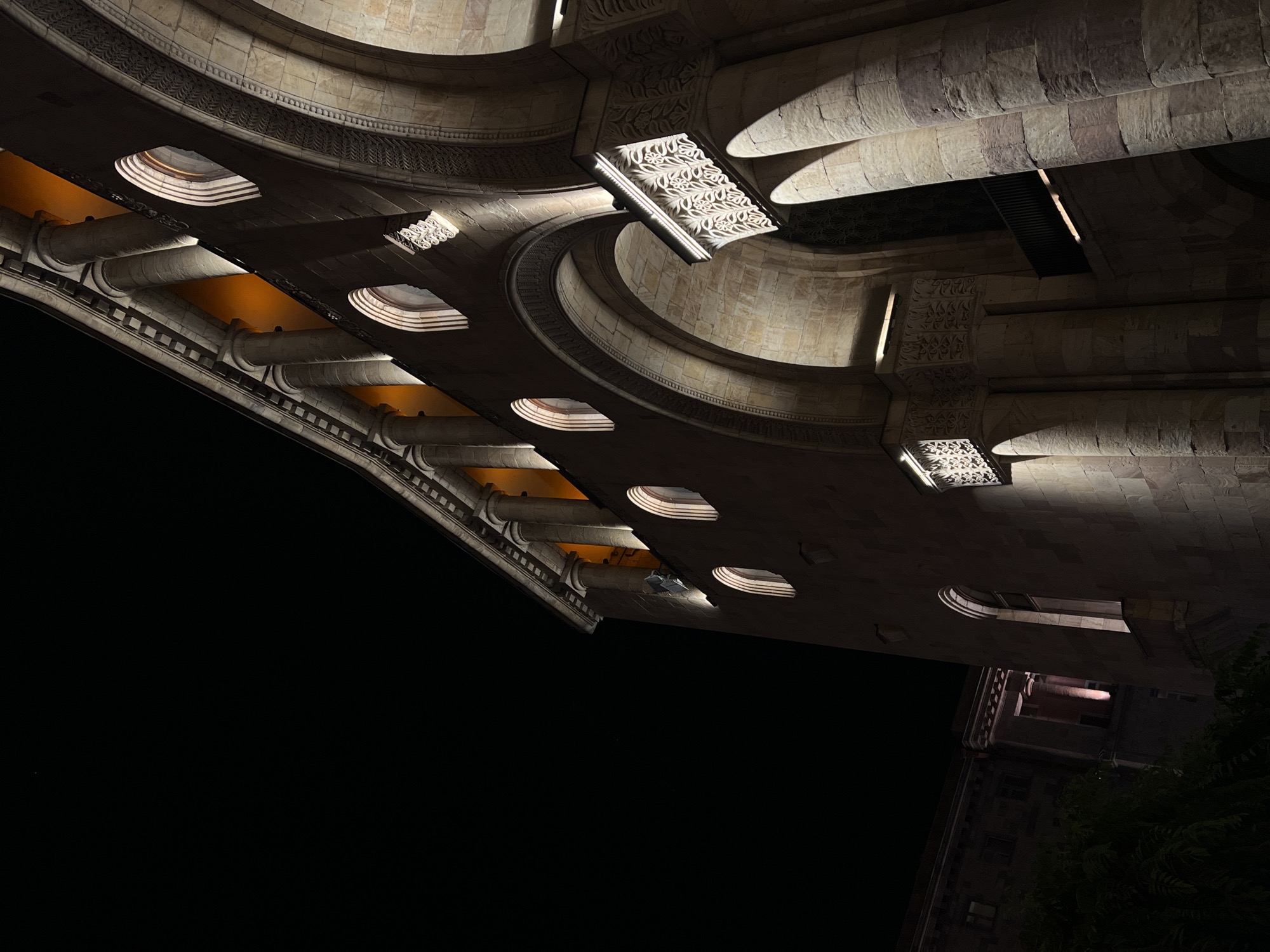 The image shows a low-angle, night-time shot of the Republic of Armenia National Academy of Sciences building in Yerevan. The building, constructed from light-colored stone, features a grand, arched entrance supported by large, round columns. Above the entrance, a colonnade of smaller columns runs along the facade. Several arched windows are visible, some illuminated from within with a warm, orange light. Intricate carvings adorn the facade, particularly around the arched openings and on decorative panels. The sky is dark, indicating it is night. To the left, a portion of another building and some dark foliage are visible, also illuminated by a soft light.