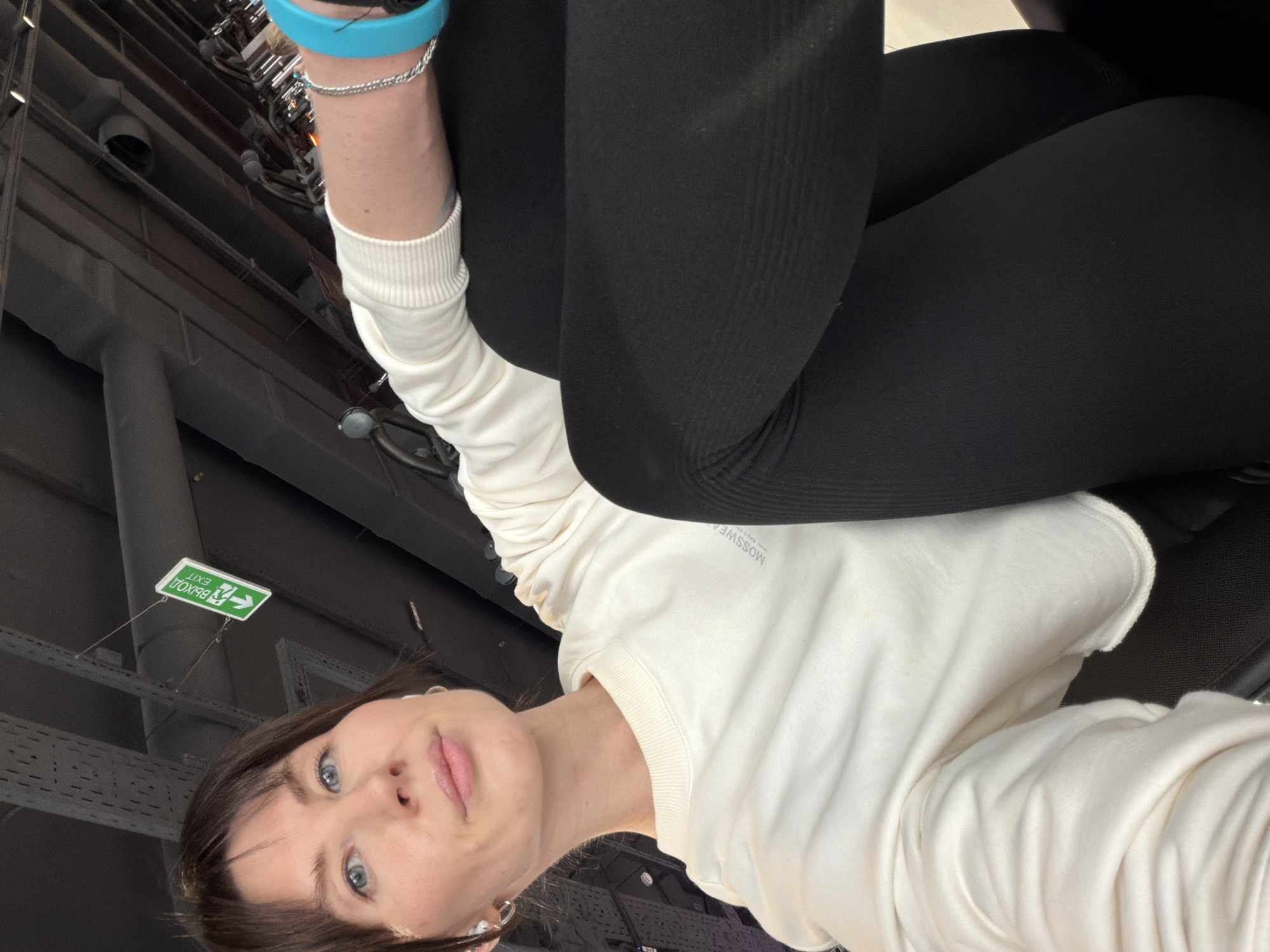 A low-angle indoor shot of a woman with fair skin and blue eyes, wearing a white sweatshirt and black leggings. She has short brown hair and is facing the camera. On her left wrist, there's a light blue wristband and a silver bracelet. Above her, an exit sign in Russian (ВЫХОД) is visible. The location appears to be an interior space in Perm, Russia. The overhead structure features gray metal supports and various utility fixtures.