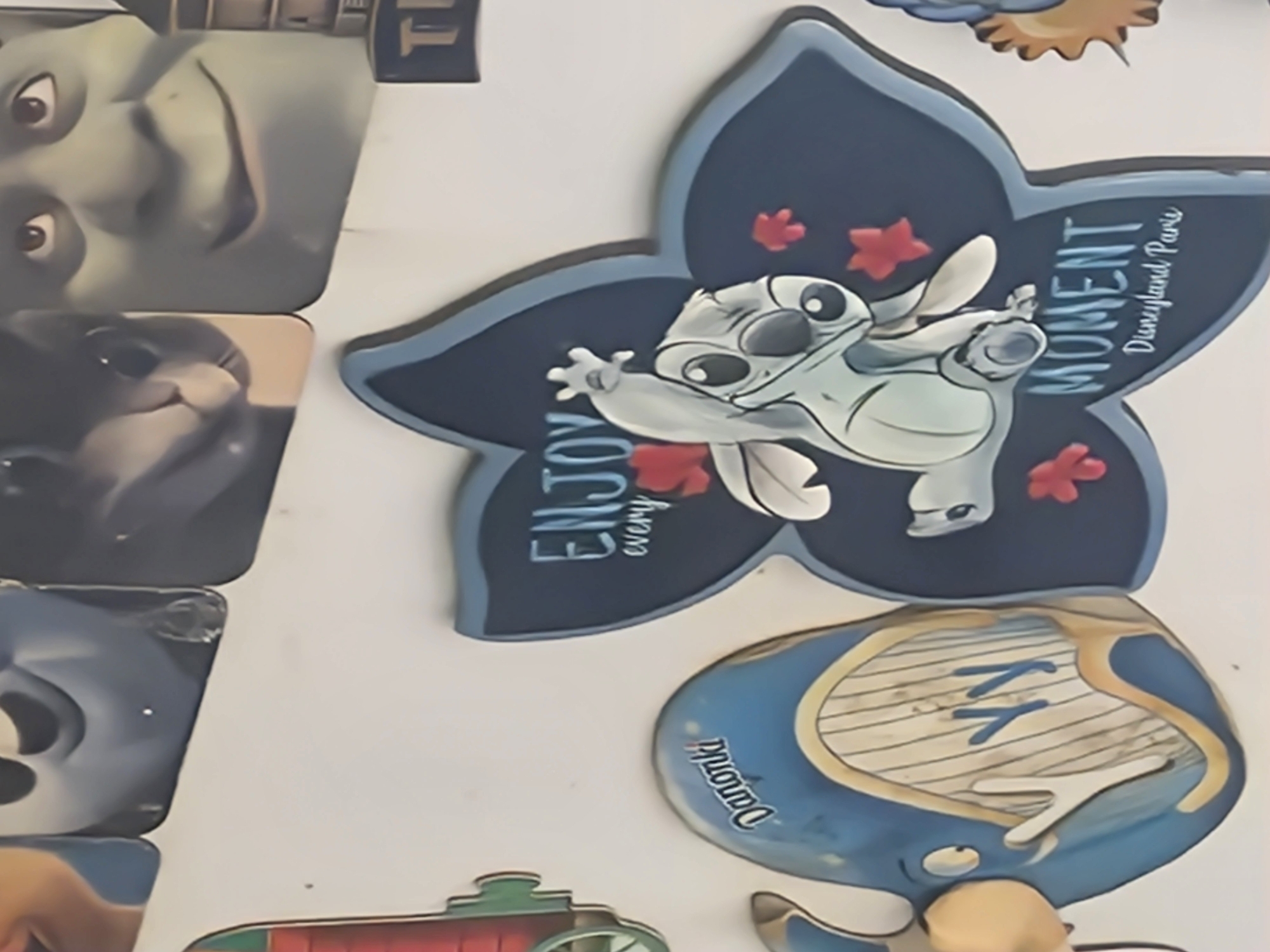 The media file displays several decorative objects, possibly magnets or wall art, against a plain white background. The dominant subject is a blue, star-shaped item featuring a cartoon character that resembles Stitch from Disney's Lilo & Stitch. The character is light blue with large ears and is depicted in a playful pose. The star shape has a dark blue outline and is adorned with small red flower-like shapes. Text within this object reads "ENJOY" and "MOMENT Disneyland Paris".
To the left of the Stitch decoration are two other decorative objects. The top one shows a close-up of a green character's face with large eyes and a wide smile, reminiscent of Shrek. The object below it features a grey cat or kitten looking curiously at the viewer. Both of these objects have rounded edges.
Further down and to the right, another decorative object is visible. It is predominantly blue and appears to be shaped like a whale or a mythical sea creature. It has yellow and white accents and contains striped patterns. Text on this object reads "Dāmu". Below this, a fragment of a red and green object with a circular detail suggests another decorative piece, possibly a toy train.
The overall impression is a collection of Disney-themed and possibly other character-based decorative items arranged together. There are no discernible clues about the time of day, weather, or specific emotions beyond the playful depiction of the characters. The location context of Ķekava, Latvia is noted but not visually evident within the media itself.