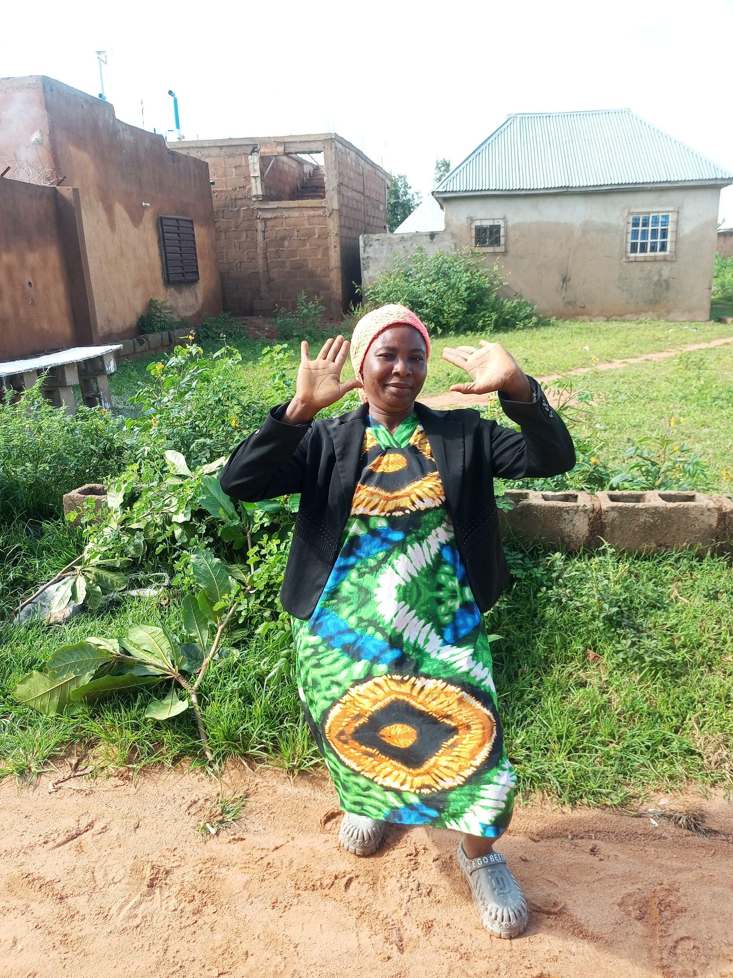 A woman with dark skin, wearing a pink headscarf, a black jacket, and a brightly colored dress with a green, blue, and orange pattern, stands in a dirt and grass area. She is facing the camera and has her hands raised to the sides of her head, with her palms facing outward. The background includes several buildings, some appearing to be under construction, with corrugated metal roofs and rough concrete or brick walls. There is also lush green vegetation and some scattered debris. The scene suggests a rural or developing urban environment, consistent with a location in Tempure, Nigeria. The lighting indicates it is daytime, with no harsh shadows suggesting midday sun.
