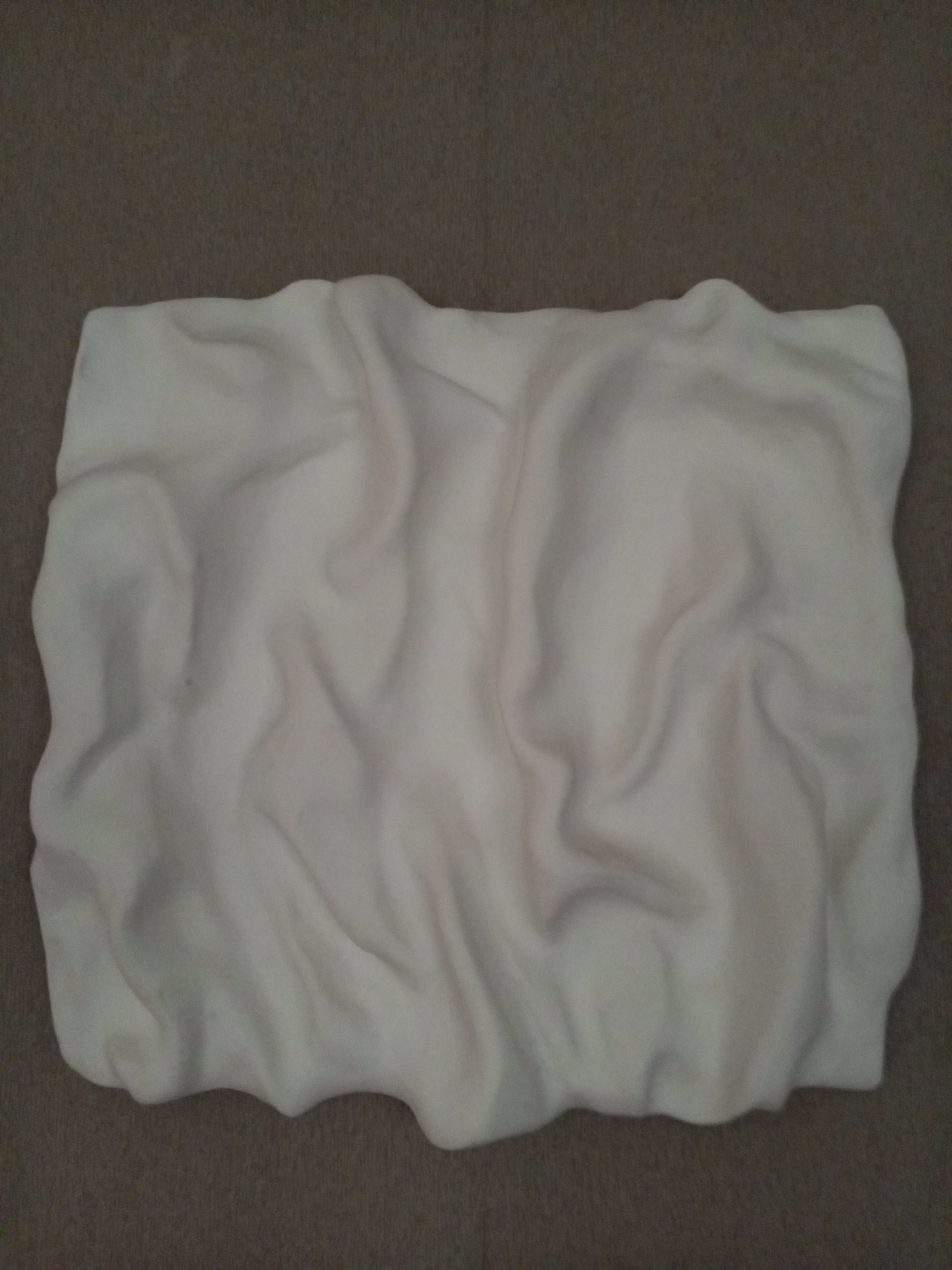 The image is a top-down, close-up shot of a folded, cream-colored fabric, likely a blanket or throw, placed on a dark, textured surface resembling carpet. The fabric is soft and has numerous wrinkles and folds, creating shadows and highlights that give it a somewhat three-dimensional appearance. There are no people or animals visible in the image. The background is uniform and out of focus. The image does not contain any discernible elements that would allow for the identification of its location within the city of Vilyuysk, Russia, or any other specific geographical location.