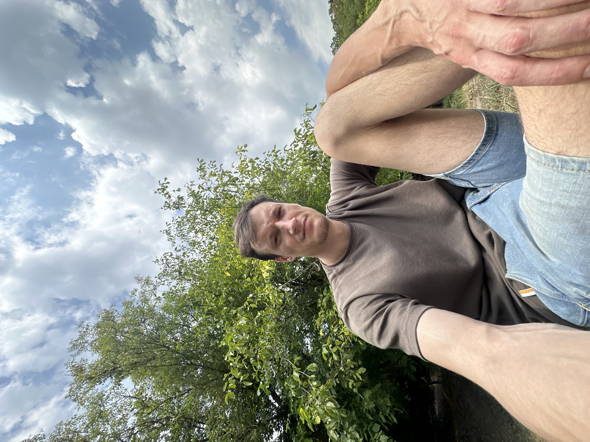 A low-angle, wide shot captures a young adult male sitting outdoors, with a leafy tree and cloudy sky as the backdrop. He is positioned in the lower half of the frame, facing the camera with a neutral expression. He wears a brown t-shirt and denim shorts. His right arm is extended forward, with his hand resting on his thigh. The sky is a mix of blue and white clouds, and the tree is lush and green. The scene appears to be in an outdoor setting, possibly a park or natural area within the city of Makiv, Ukraine.