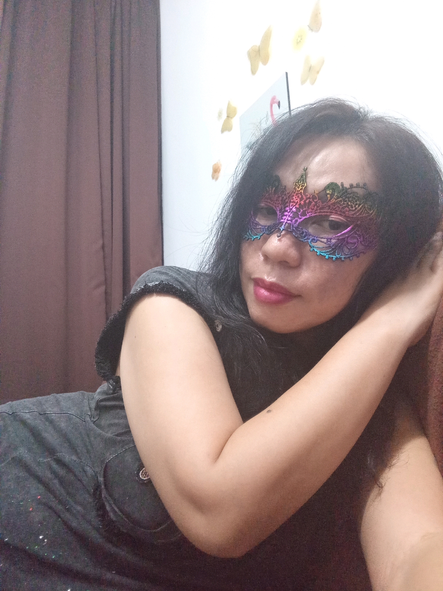 A woman, wearing a colorful masquerade mask and a dark t-shirt, lies on her side with her left arm resting on her head. Her right arm is bent, with her hand supporting her head near her temple. She is looking towards the camera with her lips slightly parted. The background consists of a brown curtain on the left and a white wall adorned with yellow butterfly decals and a picture of a flamingo on the right. The scene appears to be indoors, possibly in a bedroom, given the curtain and wall decorations. There is no discernible urban context to identify the specific location within Selayang Municipal Council, Malaysia.