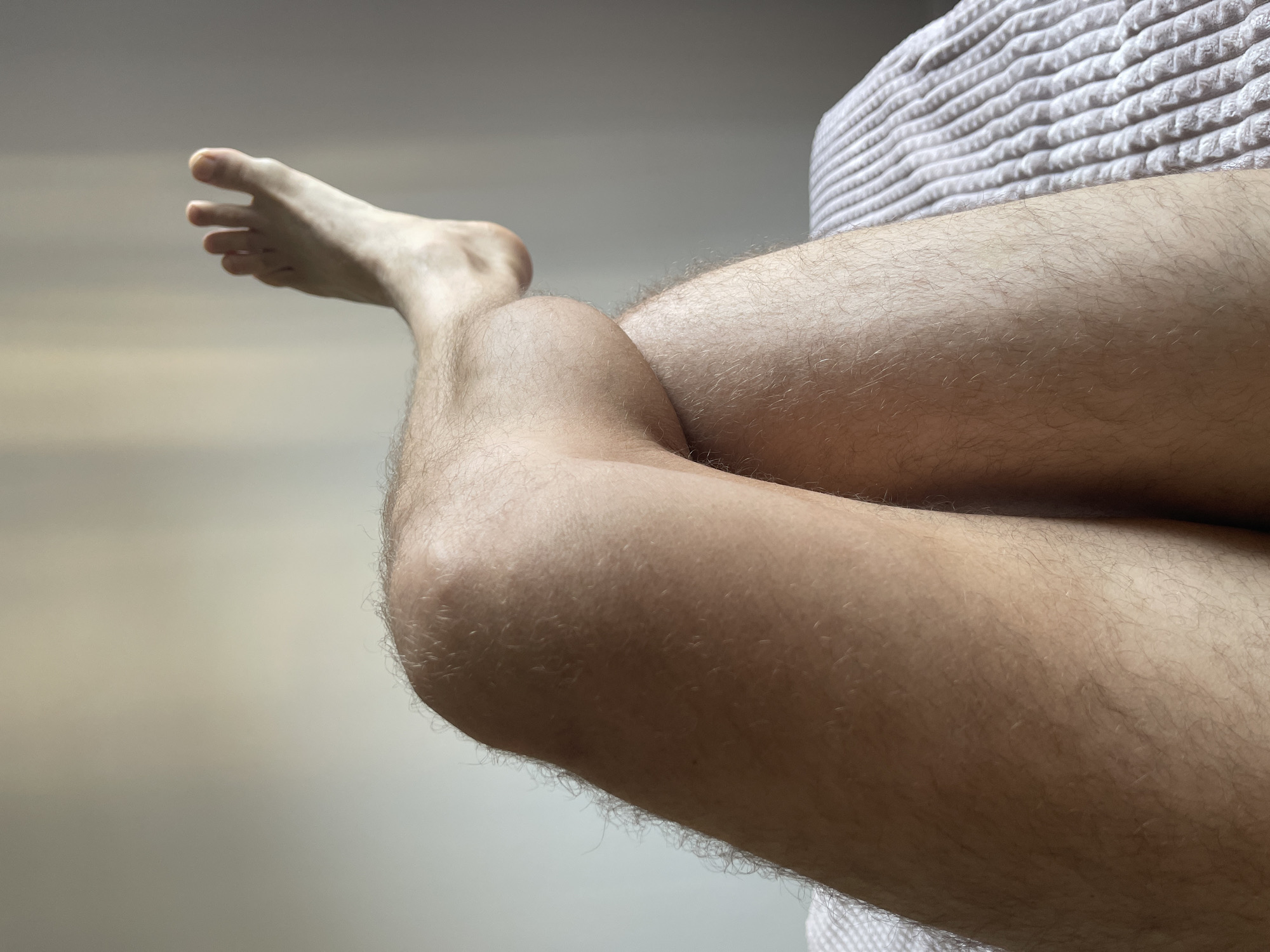 The image shows a close-up view of a person's legs resting on a white textured surface, possibly a blanket or cushion. The legs are fair-skinned with light-colored body hair. The foot is extended with toes visible. The background is a blurry, neutral-toned wall. The location is not evident from the image, and there is no indication of Izhevsk, Russia.