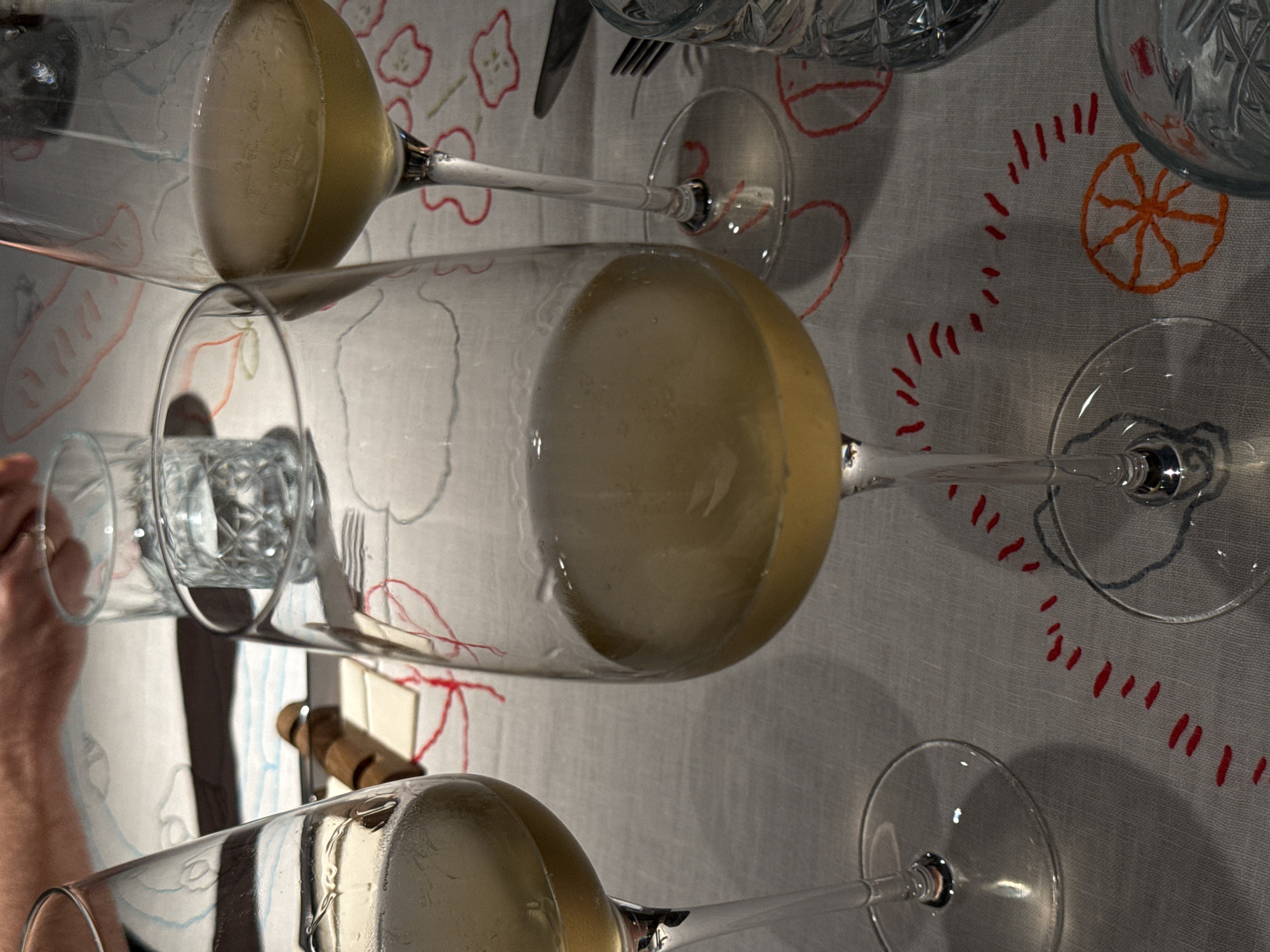 The image captures an indoor dining table scene in Vracar Urban Municipality, Serbia. Three clear stemmed glasses, each containing a pale yellow liquid consistent with white wine, are positioned on a light-colored linen tablecloth. The tablecloth is embroidered with a pattern of red dashed lines, abstract red outlines, and an orange, spoked circular design. A central wine glass is prominently displayed, largely full. A second wine glass is partially visible on the far left, also containing liquid, and a third is partially visible on the right, containing a lesser amount of liquid. Two clear, cut-glass tumblers are present: one in the mid-background and another partially visible in the lower right. In the upper background, human hands are visible, one resting on the tablecloth near a horizontally placed knife with a wooden handle and several small, light-colored rectangular items. A silver fork is partially visible to the right of the central wine glass. Shadows from the glassware are cast onto the tablecloth, indicating an overhead light source.
