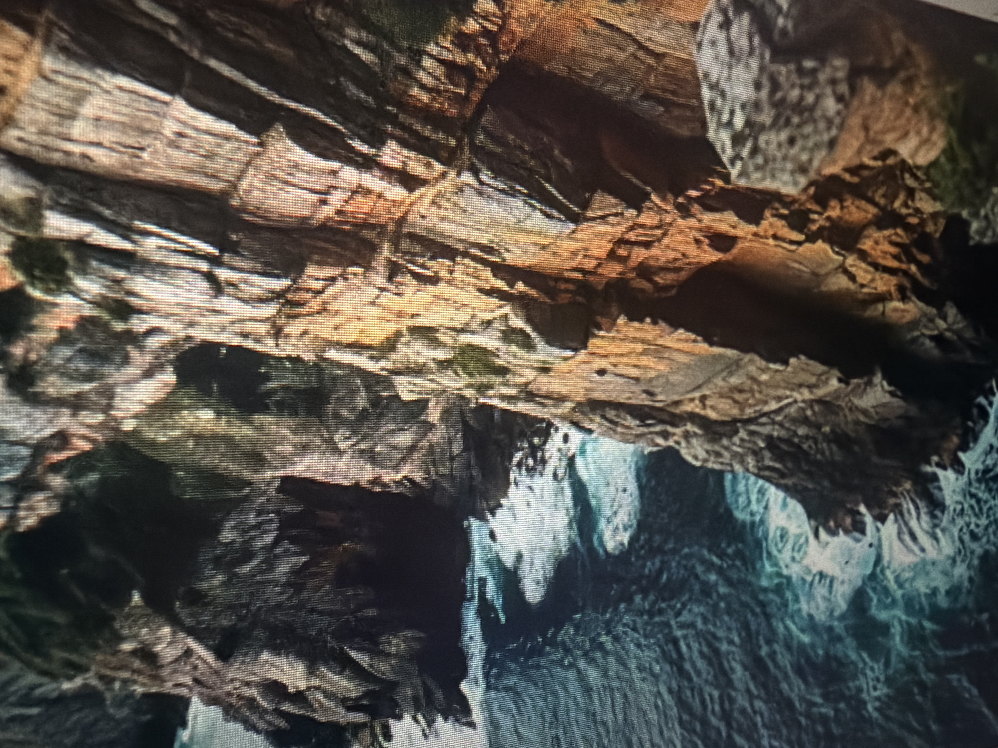 The image shows a rugged, rocky coastline with the ocean crashing against the cliffs. The cliffs are sheer and appear to be made of sedimentary rock, with layers visible. The water is a deep blue-green, and there are whitecaps and waves breaking near the shore. The scene is dimly lit, with shadows obscuring some of the details of the cliffs and the water. There is a dark opening or cave visible in the cliff face on the left side of the image. The overall impression is one of a wild and untamed natural landscape. There is no indication of the city of Baan Mae Nam, Thailand, in the image.