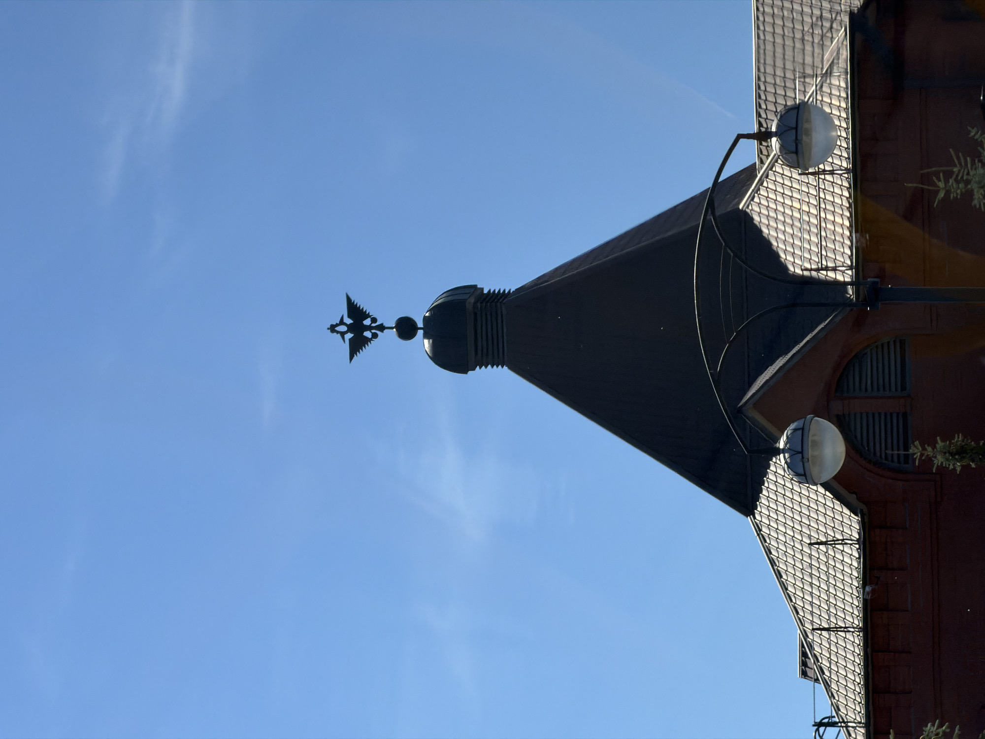 The image shows the top of a building with a dark conical roof and a pitched roof covered in shingles. A black metal double-headed eagle is perched on top of the conical roof. Two globe-shaped streetlights with curved metal arms are visible in front of the pitched roof. The building appears to be constructed of red brick and has arched windows with louvers. The sky is clear and blue with a few faint wisps of clouds. The image is taken from a low angle, looking up at the building.
