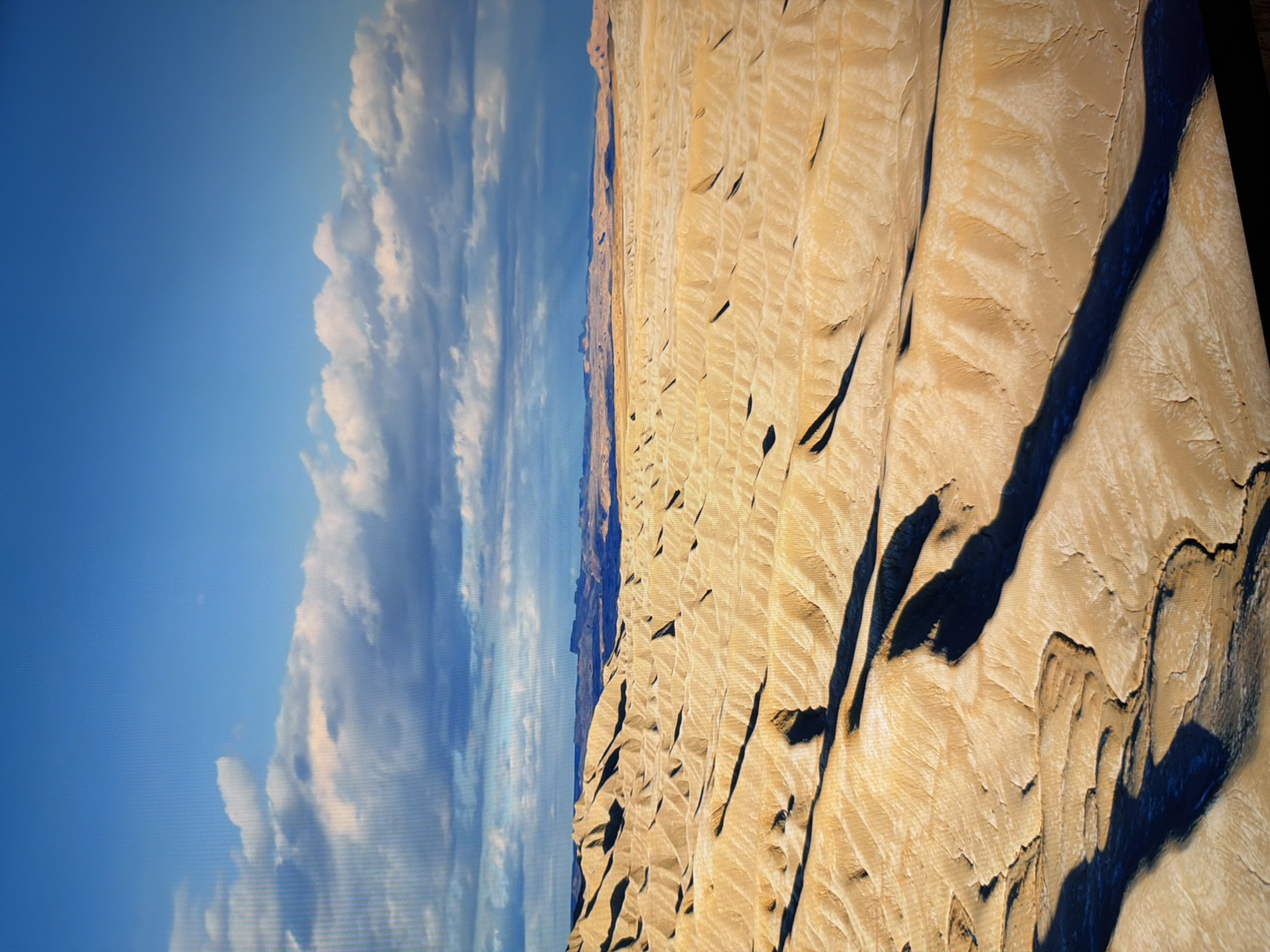 The image depicts a vast, arid desert landscape under a bright blue sky with scattered white and grey clouds. The terrain is characterized by undulating sand dunes and eroded rock formations that cast long, dark shadows. In the distance, a hazy horizon line suggests more distant mountains or geological features. The colors are dominated by warm yellows and browns of the desert, contrasted with the vibrant blue of the sky. There is no indication of any urban environment, including the city of Kyiv, Ukraine, nor are there any people, subjects, objects, or actions depicted. The scene is a natural landscape.