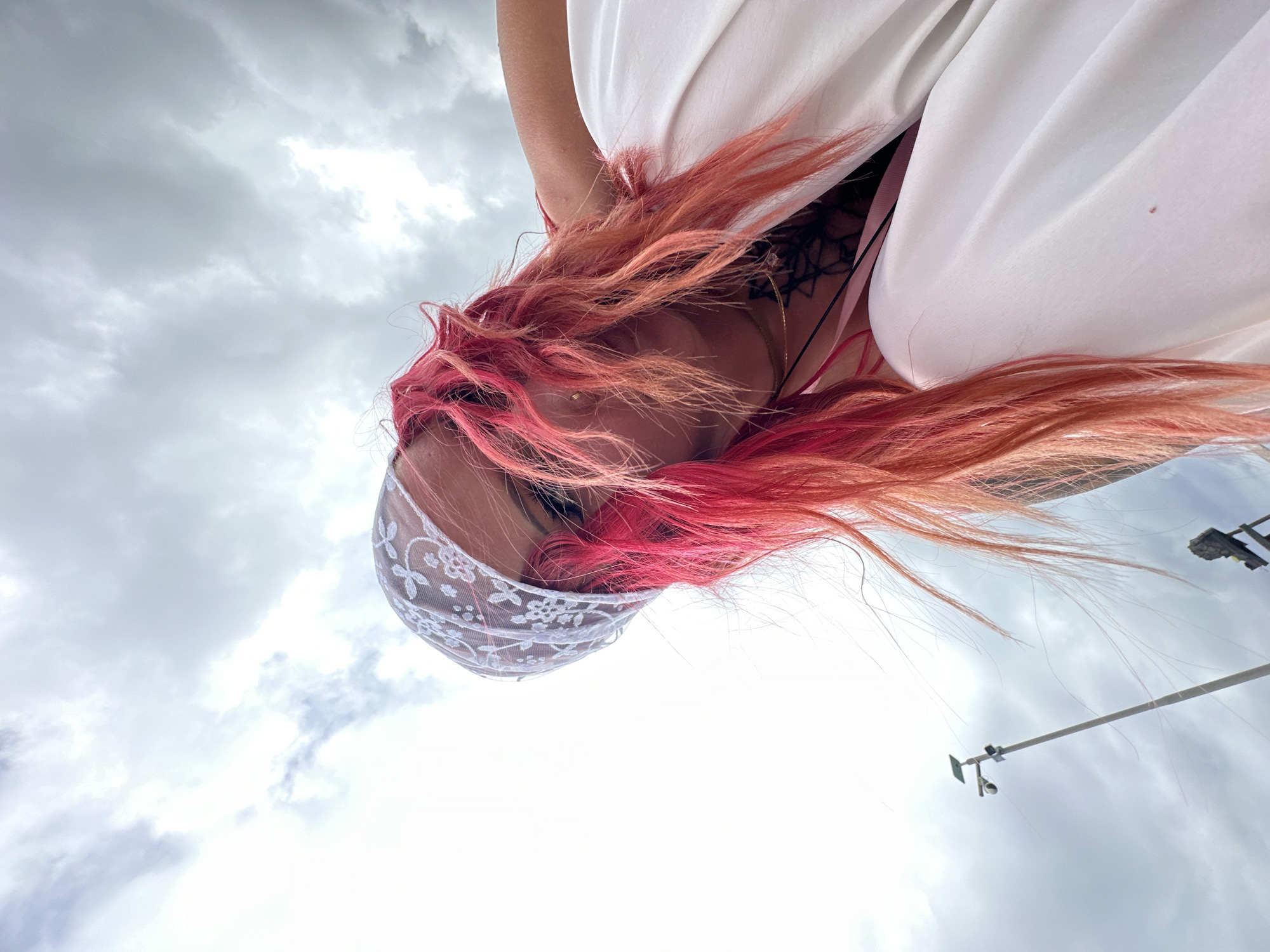 A low-angle shot depicts a person with vibrant pink and red hair wearing a white lace head covering. The person is looking down, and a white garment obscures much of the view. A black tattoo is visible on the upper chest. The backdrop features a cloudy sky and a lamppost. The location is Kampung Toyapakeh, Indonesia.