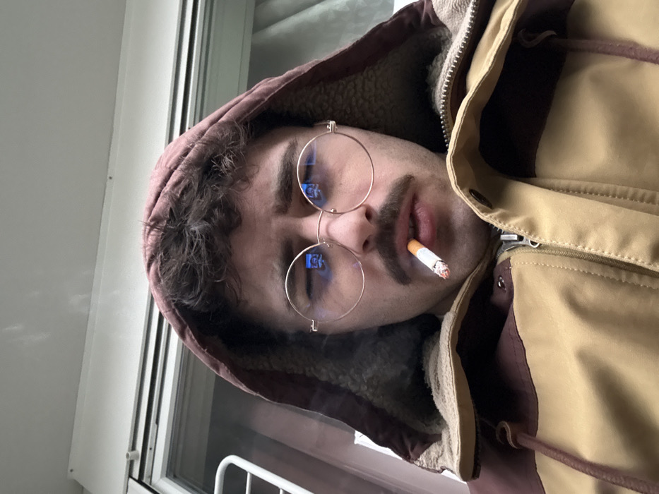 A man with fair skin, a dark mustache, and curly hair wears round glasses and a brown hooded jacket with a tan shoulder area. He holds a lit cigarette between his lips, smoke visible around it. Part of a white window frame is visible in the background, near the lower left. The image is slightly rotated clockwise.