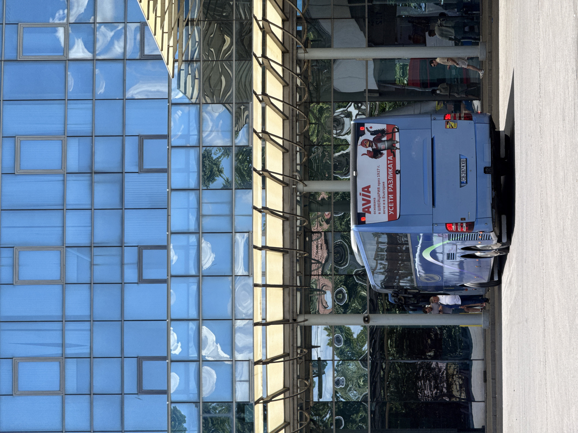 The image shows the rear of a blue bus parked in front of a modern glass-facade building. The bus has an advertisement on its rear that reads "AVIA" and "УСЕТИ РАЗЛИКАТА" in Cyrillic script, indicating the location is likely in Bulgaria, and specifically Sofia based on the license plate format which appears to be a Bulgarian one. The building's glass panels reflect the sky, clouds, and surrounding trees, creating a distorted and abstract pattern. There are several people standing near the bus and the entrance of the building. One person is leaning against a pillar while looking at their phone, and another is walking away from the bus. The environment appears to be an urban setting, possibly a bus station or a public transportation hub. The time of day is daytime, with bright sunlight casting shadows.